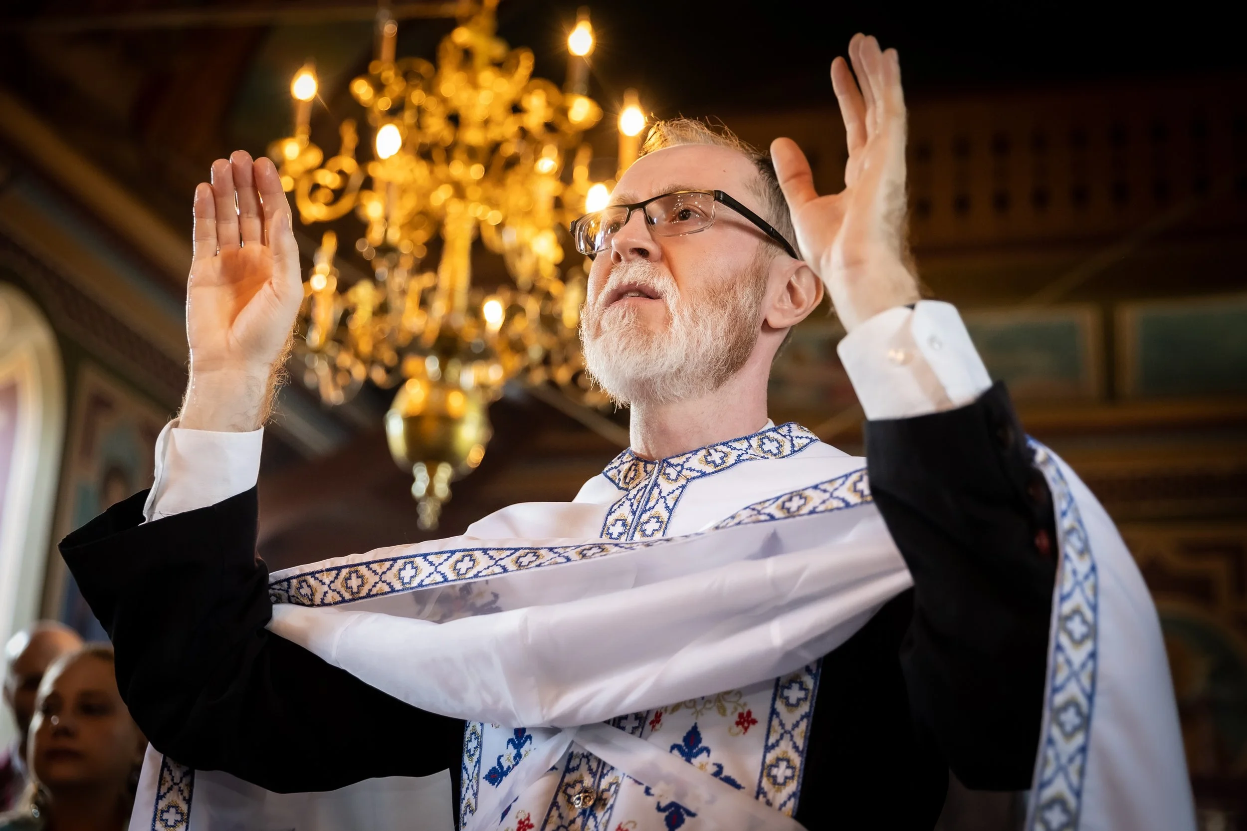 A man wearing glasses and traditional embroidered clothing is performing a religious or cultural ritual, raising both hands with palms facing outward inside a church or cathedral with a chandelier in the background.