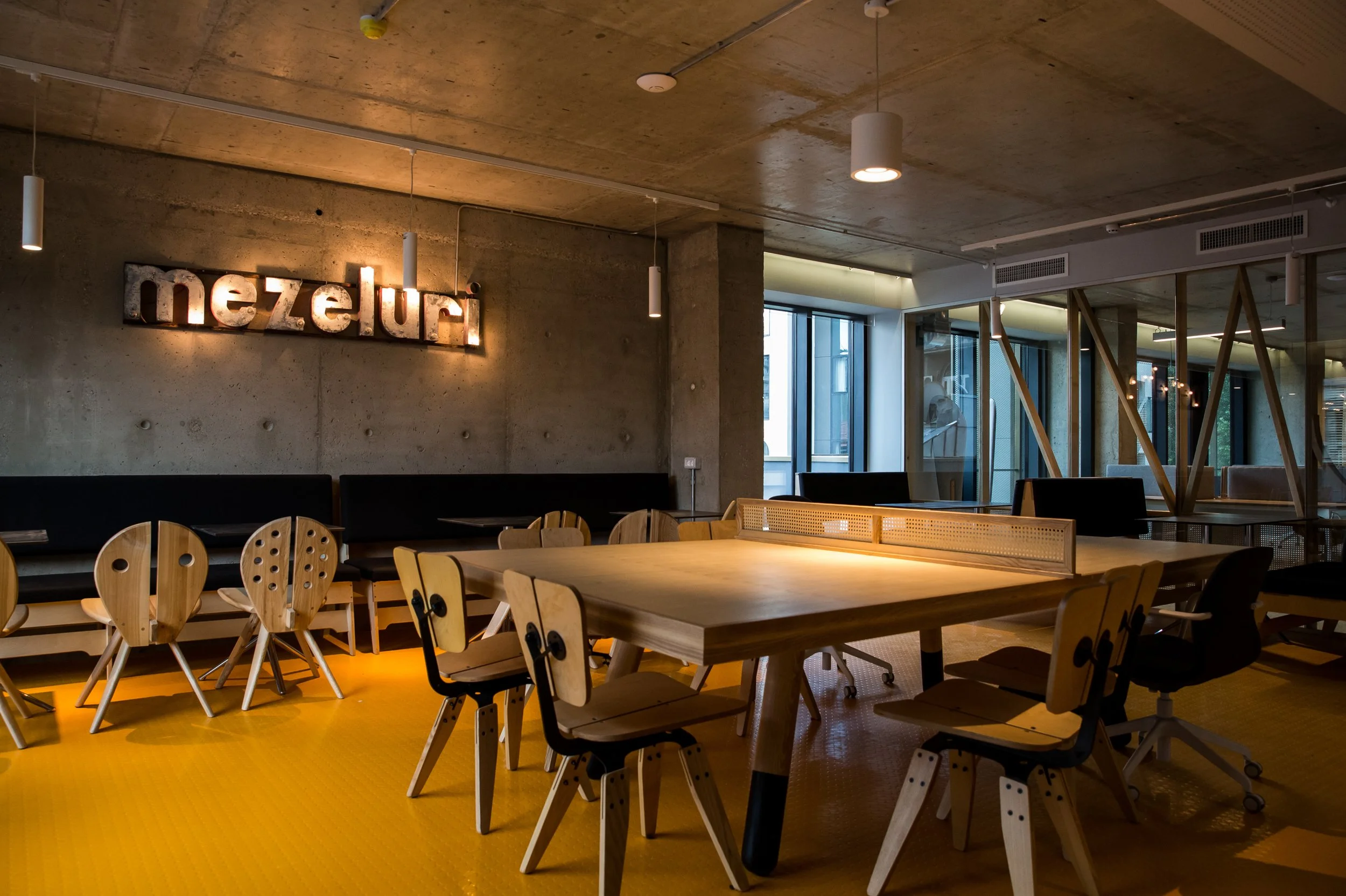 Interior view of a modern office or co-working space with wooden tables, chairs, and a concrete ceiling. There is a neon sign on the wall that reads 'nezu', and large windows letting in natural light.