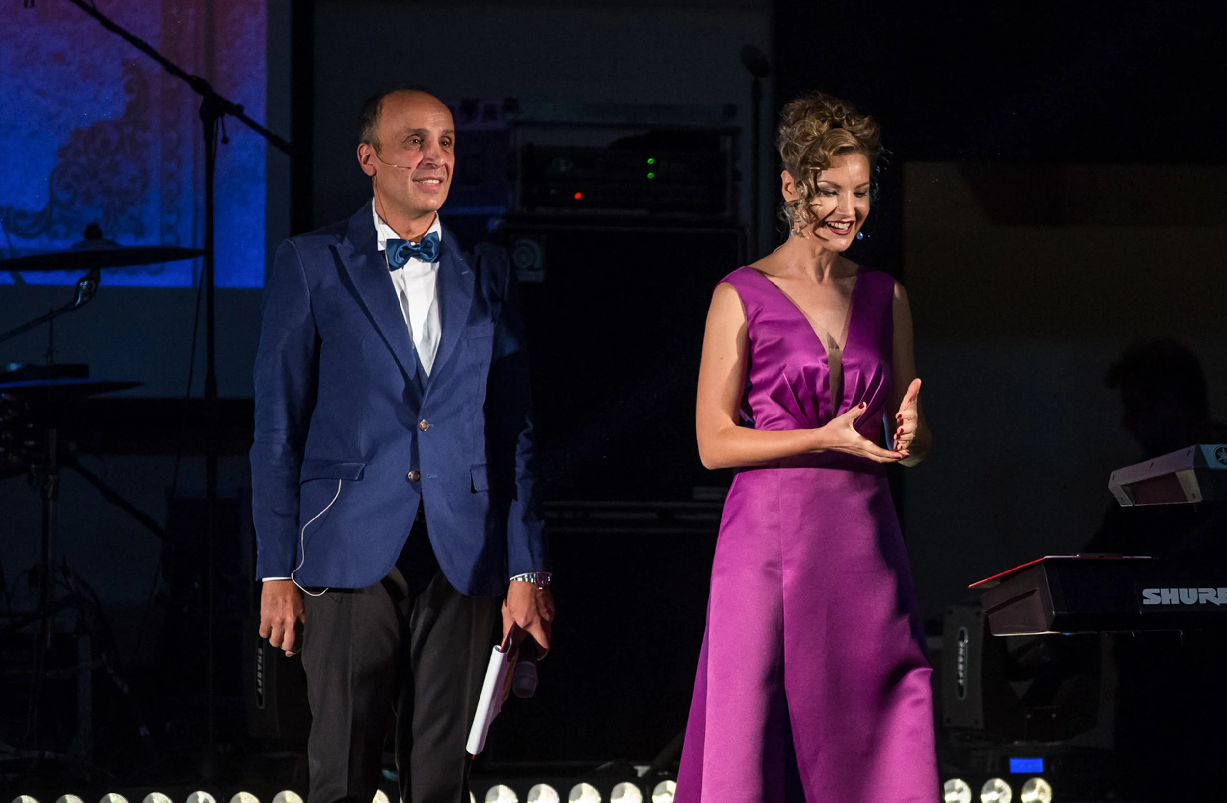 A man and woman are on stage during a performance or presentation. The man is dressed in a blue suit with a bow tie, and the woman is wearing a purple dress. They are smiling, and the woman appears to be speaking or presenting, holding a paper or car