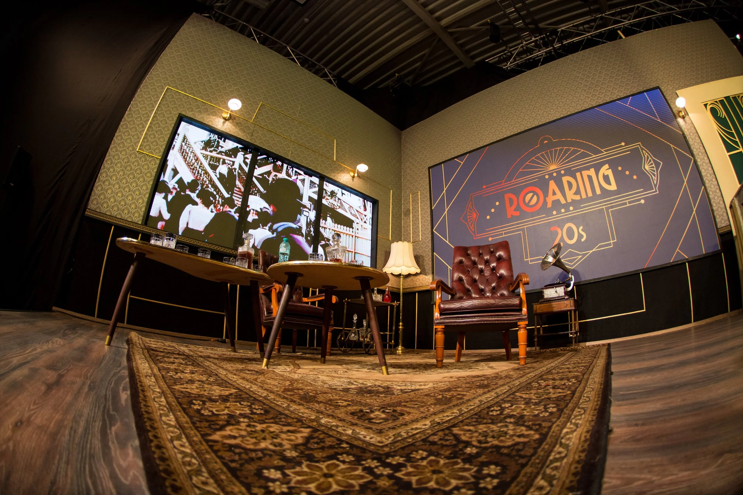An indoor venue with a vintage style, featuring a patterned area rug, two brown leather armchairs, a table with drinks, a floor lamp, a large screen displaying an image, and a wall with geometric designs and the word 'ROARING' in bold letters.