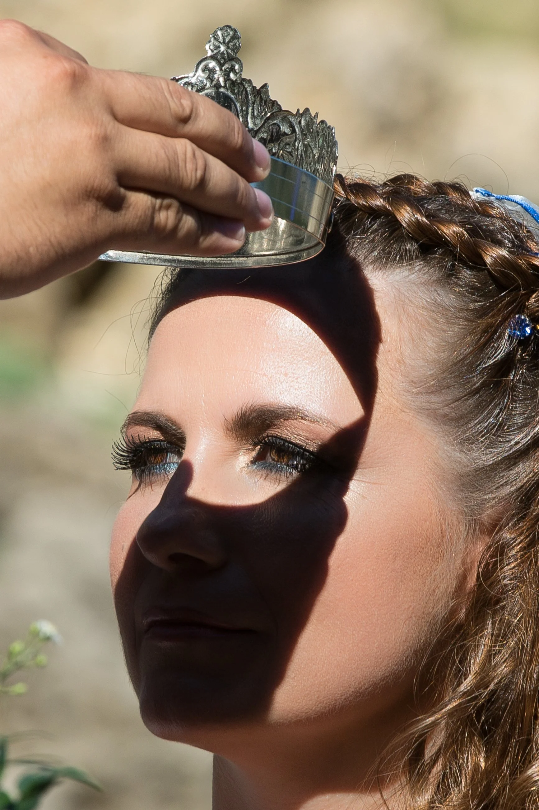 A woman with braided hair and makeup is receiving a crown placed on her head during a outdoor event, with a blurred natural background.