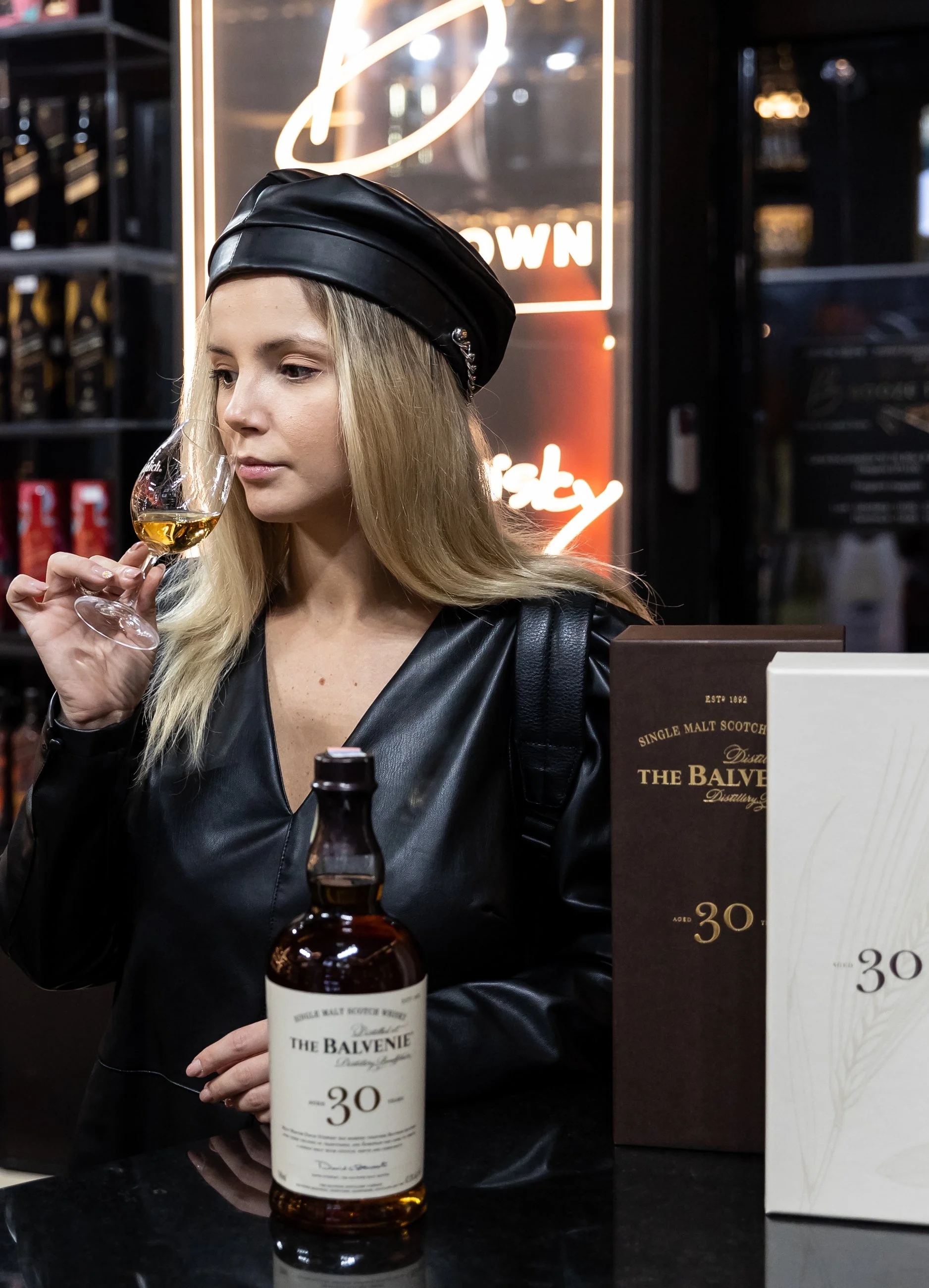 Young woman with blonde hair wearing a black leather hat and leather jacket, holding a glass of whiskey with a thoughtful expression, in a bar or liquor store with bottles in the background and a whiskey box on the counter.