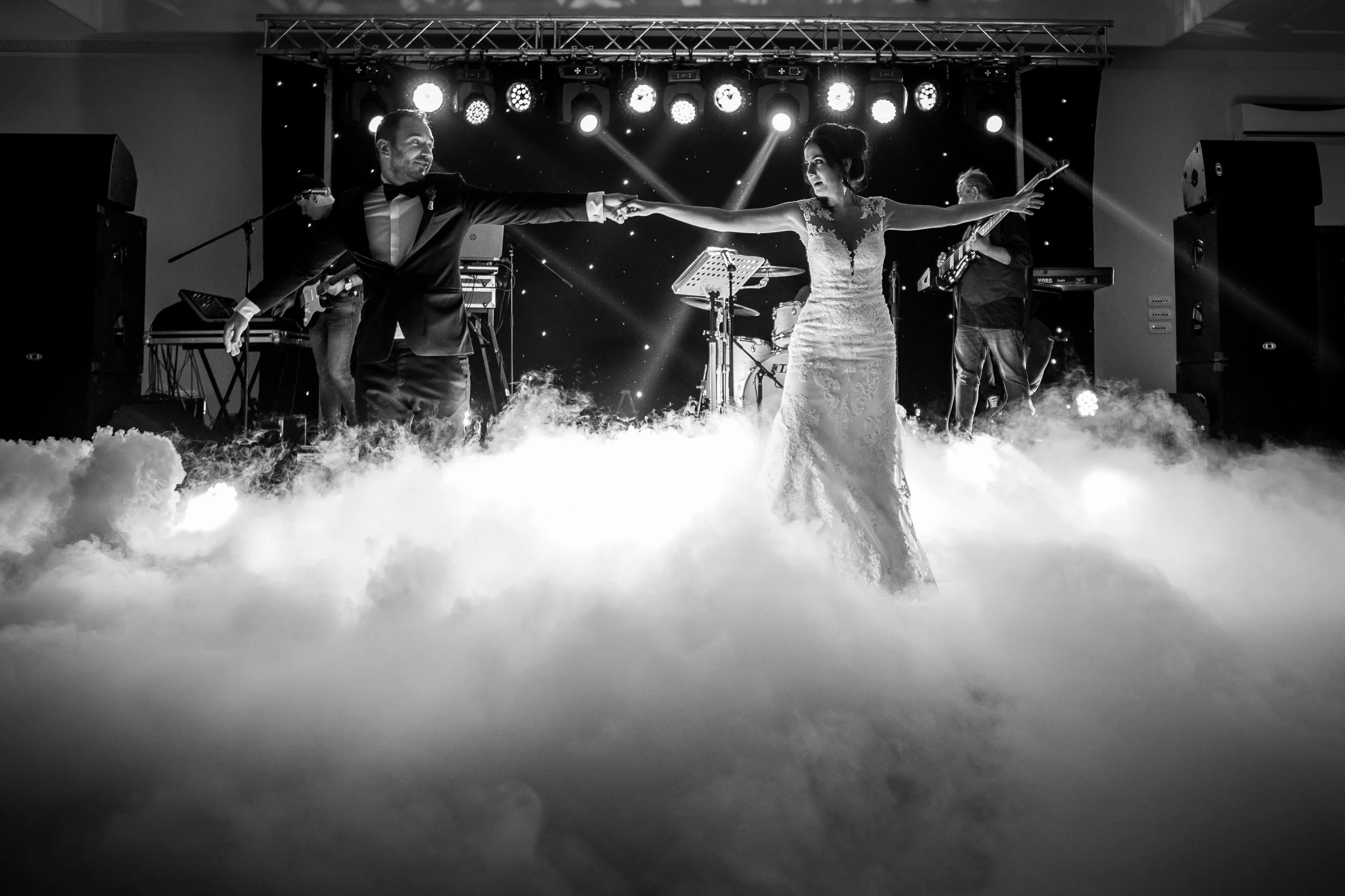 Black and white photo of a bride and groom dancing on a stage with bright lights and fog at a wedding reception.