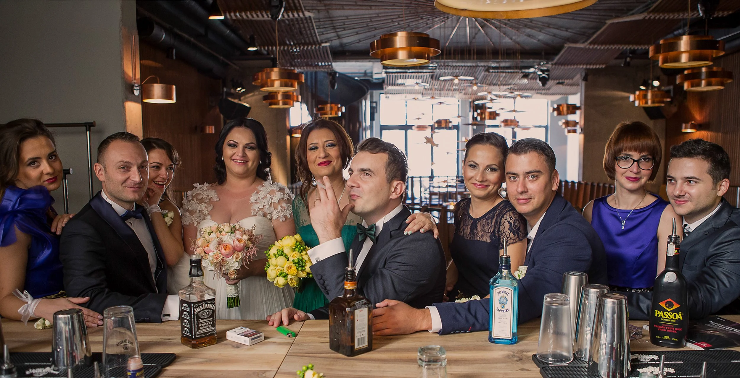 A group of nine people, including a bride and groom, gathered around a table during a celebration in a restaurant or bar with modern decor and warm lighting.