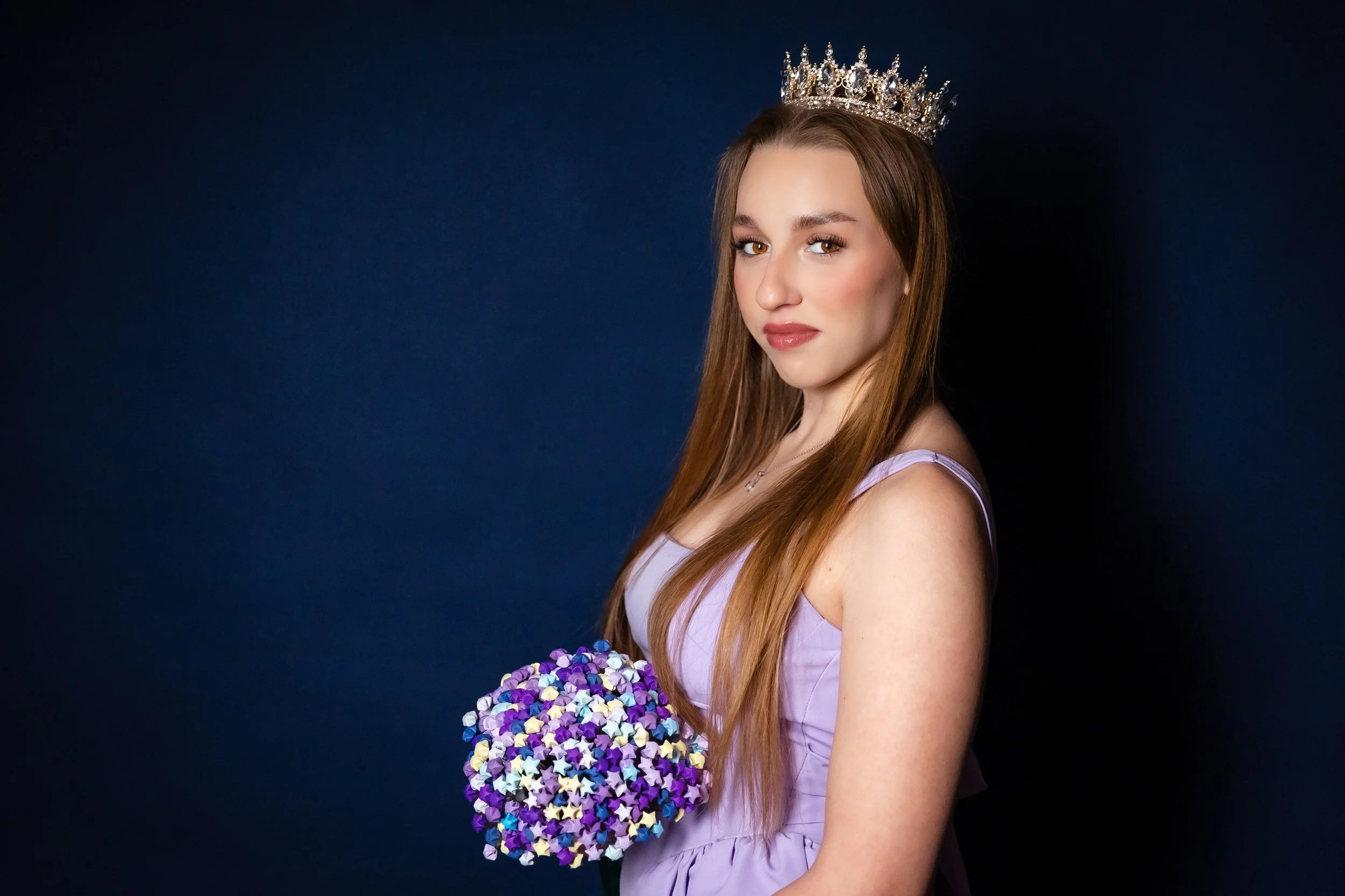 Young woman wearing a silver crown, holding a bouquet of multicolored star-shaped candies, dressed in lavender with a dark background.