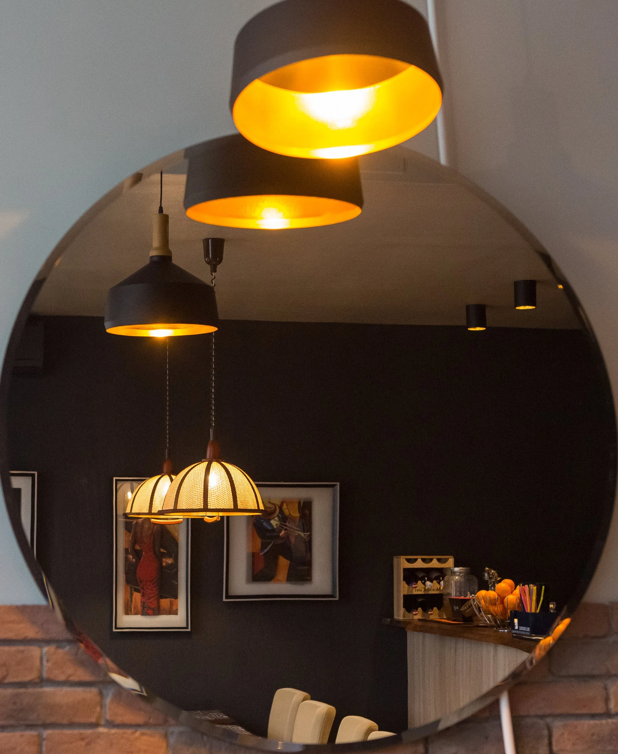 Interior scene reflected in a mirror showing hanging pendant lights with black and gold shades, a framed artwork, a small shelving unit with knickknacks, and a wooden countertop with various items including oranges and stationery, against a dark wall