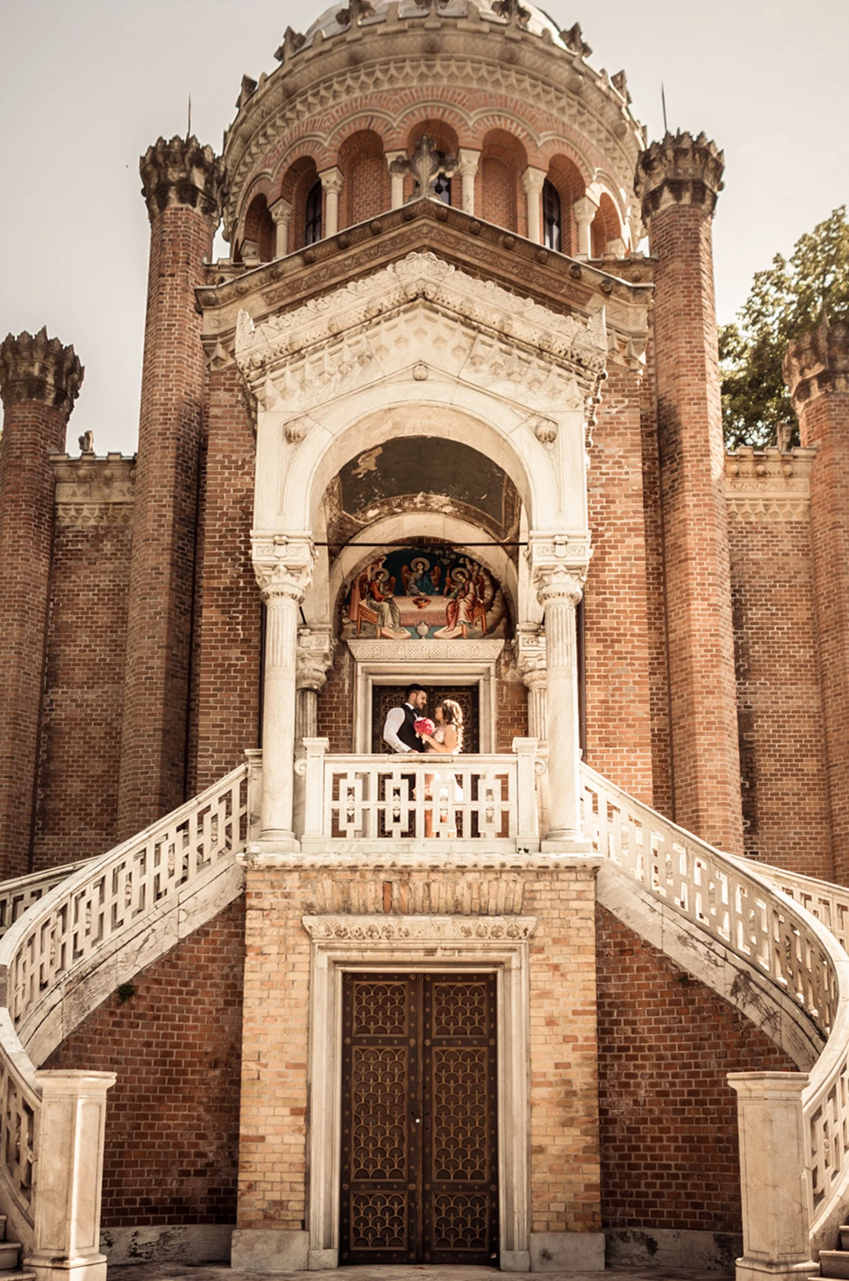 A couple stands on a staircase in front of a historic brick building with ornate architecture and religious murals.