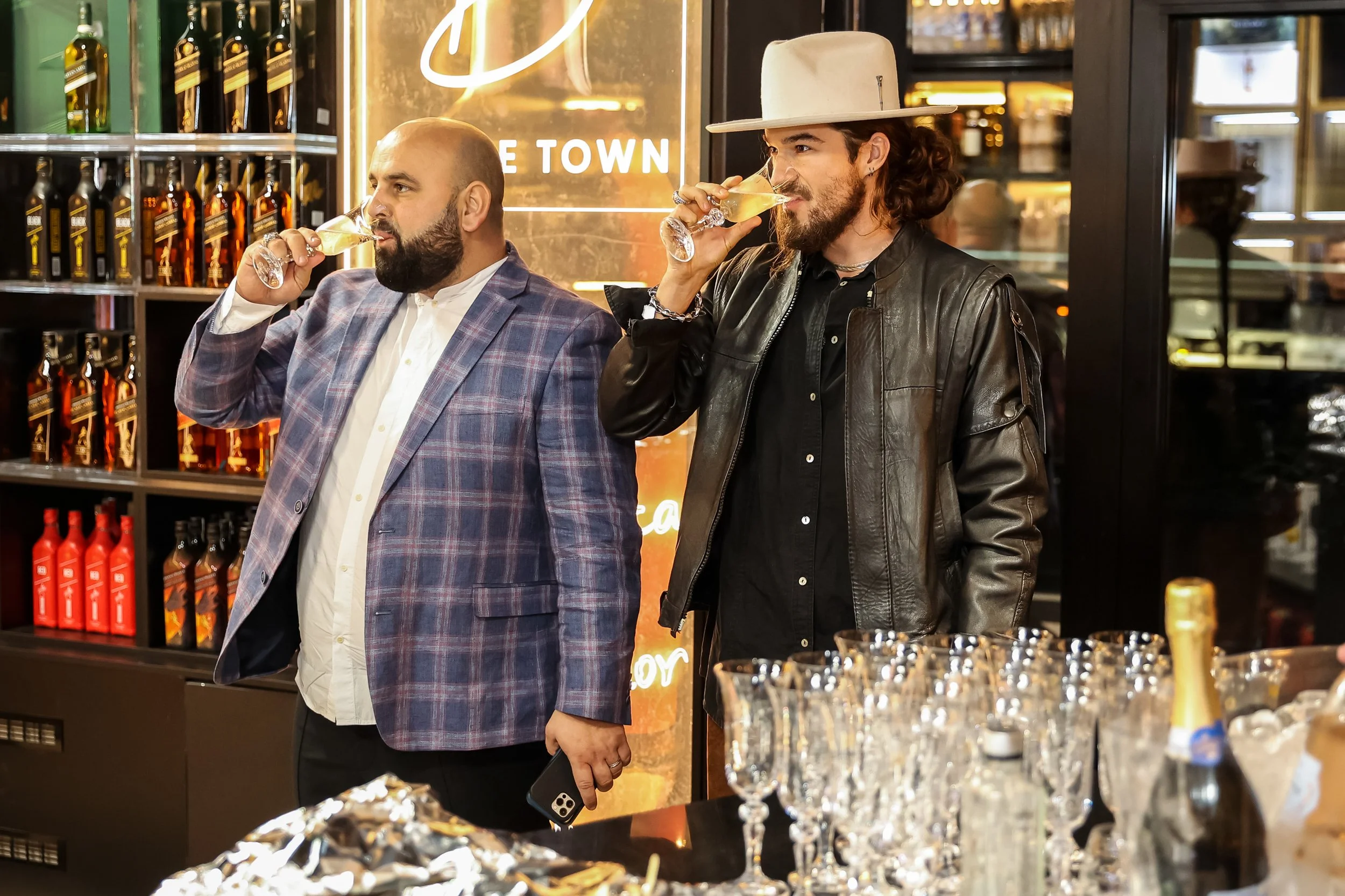 Two men are standing at a bar, sipping champagne. The man on the left is wearing a checkered blazer and has a beard, while the man on the right has long hair, a hat, and is wearing a leather jacket. There are shelves of liquor bottles in the backgrou