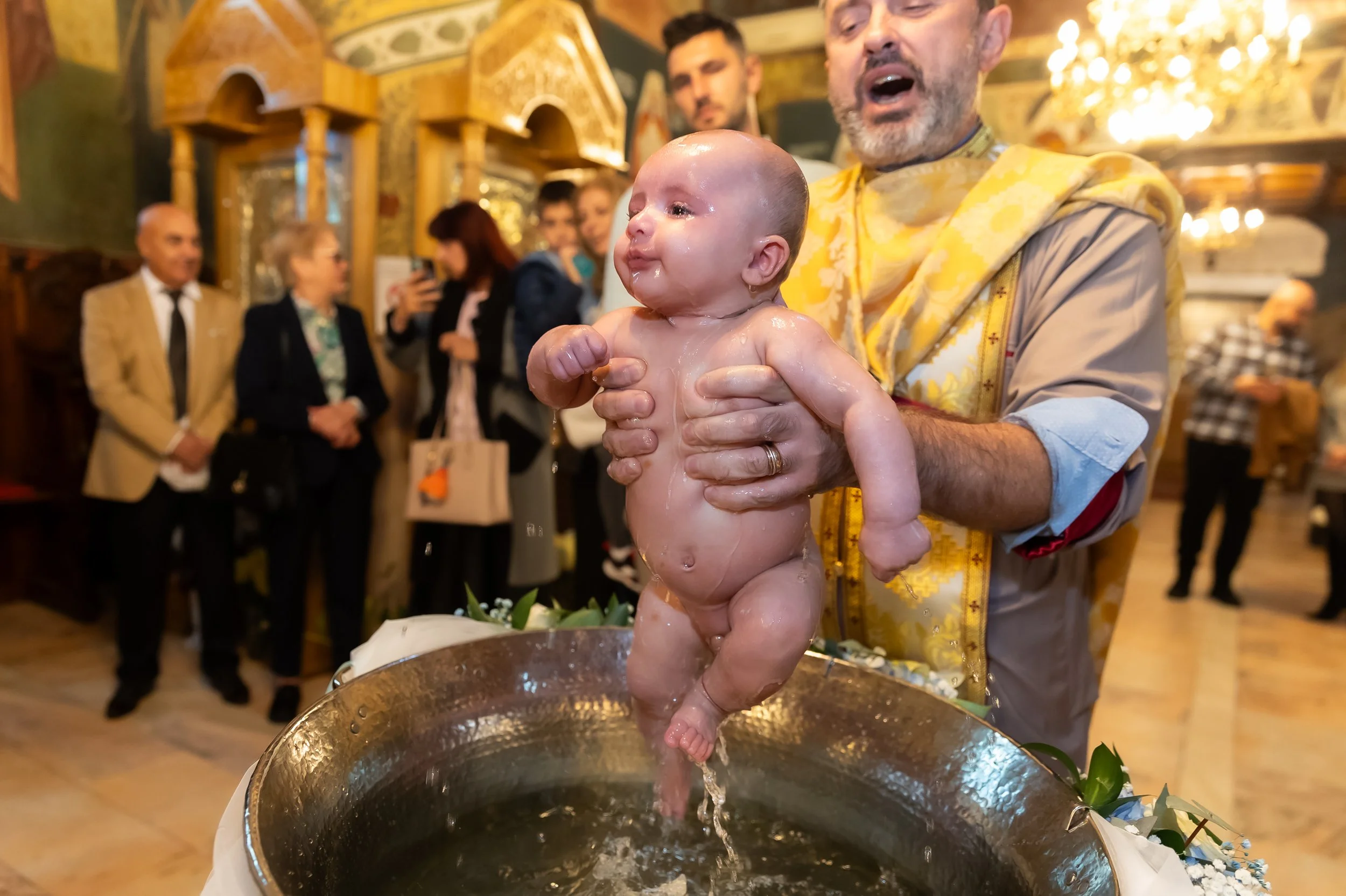 A baby being baptized with water poured over its head by a man at a church or religious ceremony, with several onlookers in the background.