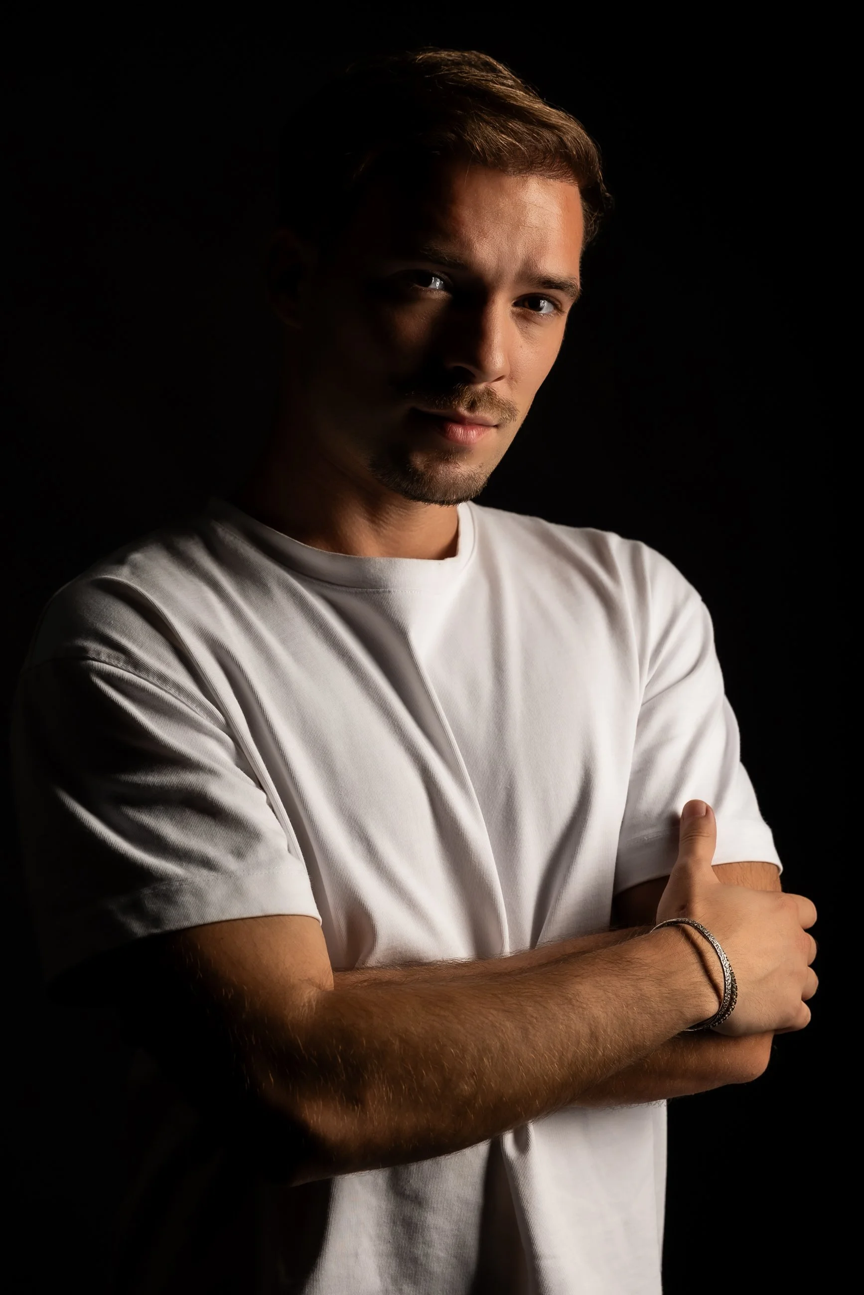 Portrait of a young man with brown hair, light facial hair, wearing a white t-shirt, posing with arms crossed against a black background.