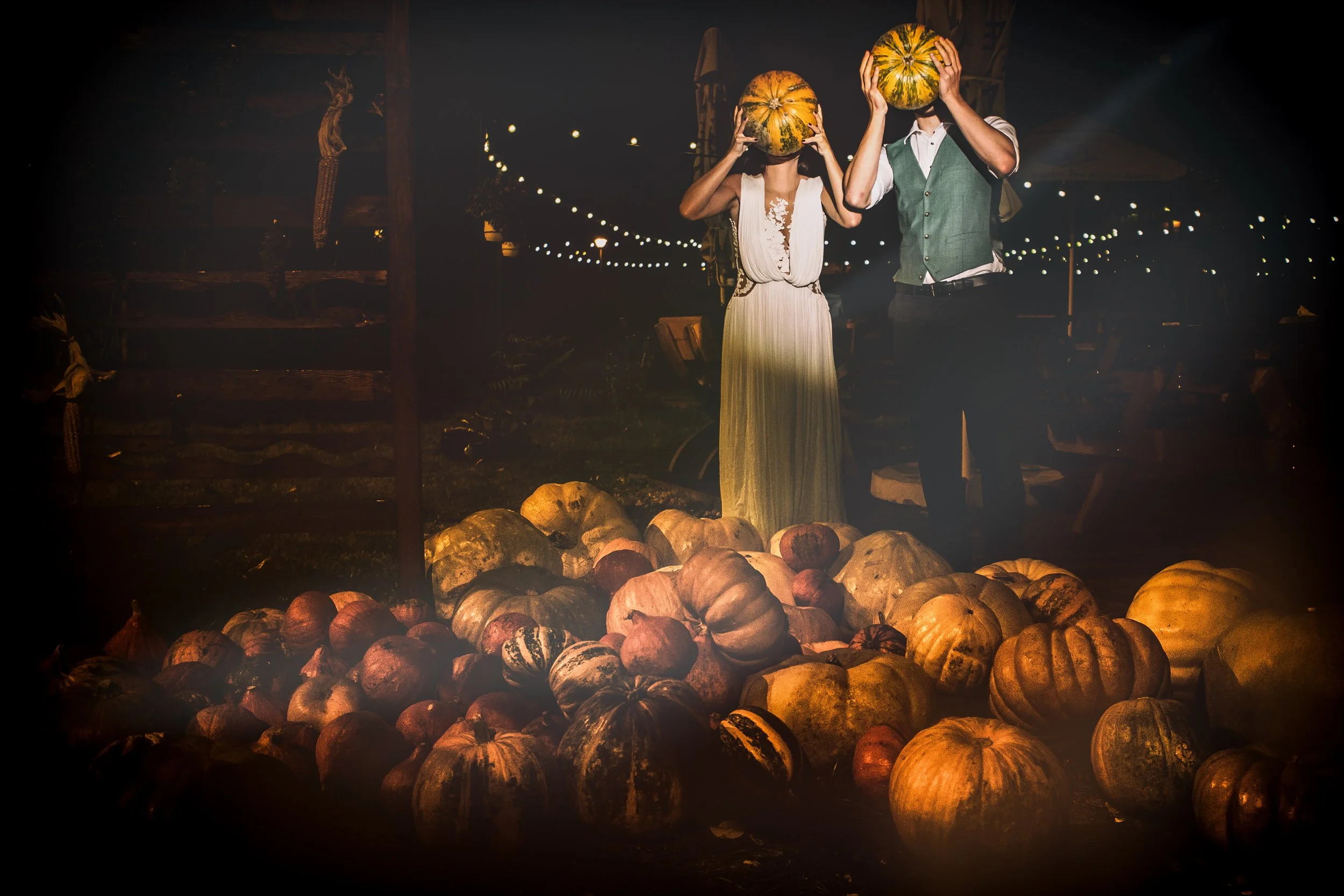Two people with pumpkins over their heads standing among pumpkins at night with string lights overhead.