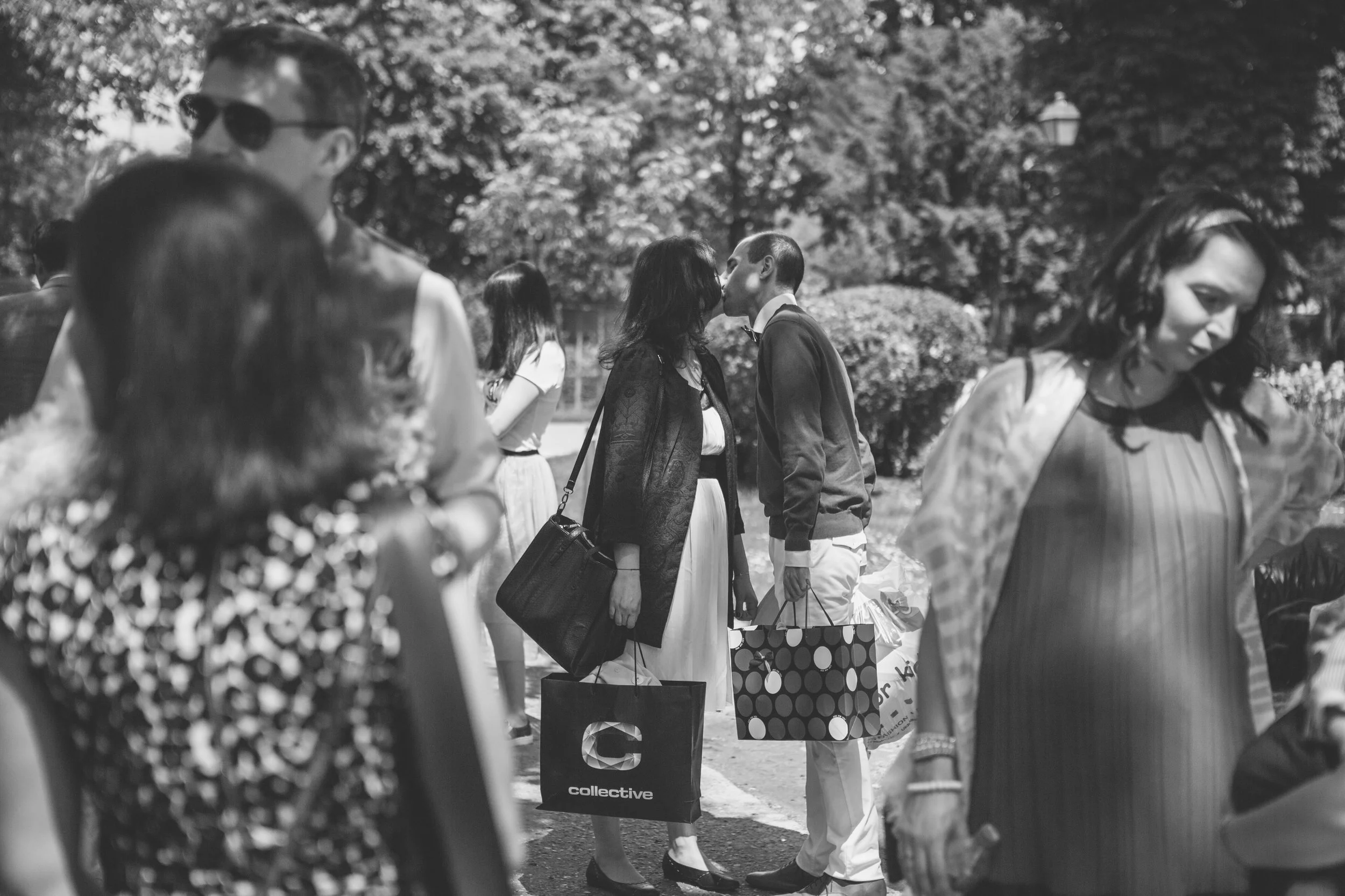 A black and white photo of a couple kissing at an outdoor event, surrounded by other people carrying shopping bags, with trees and a lamp post in the background.