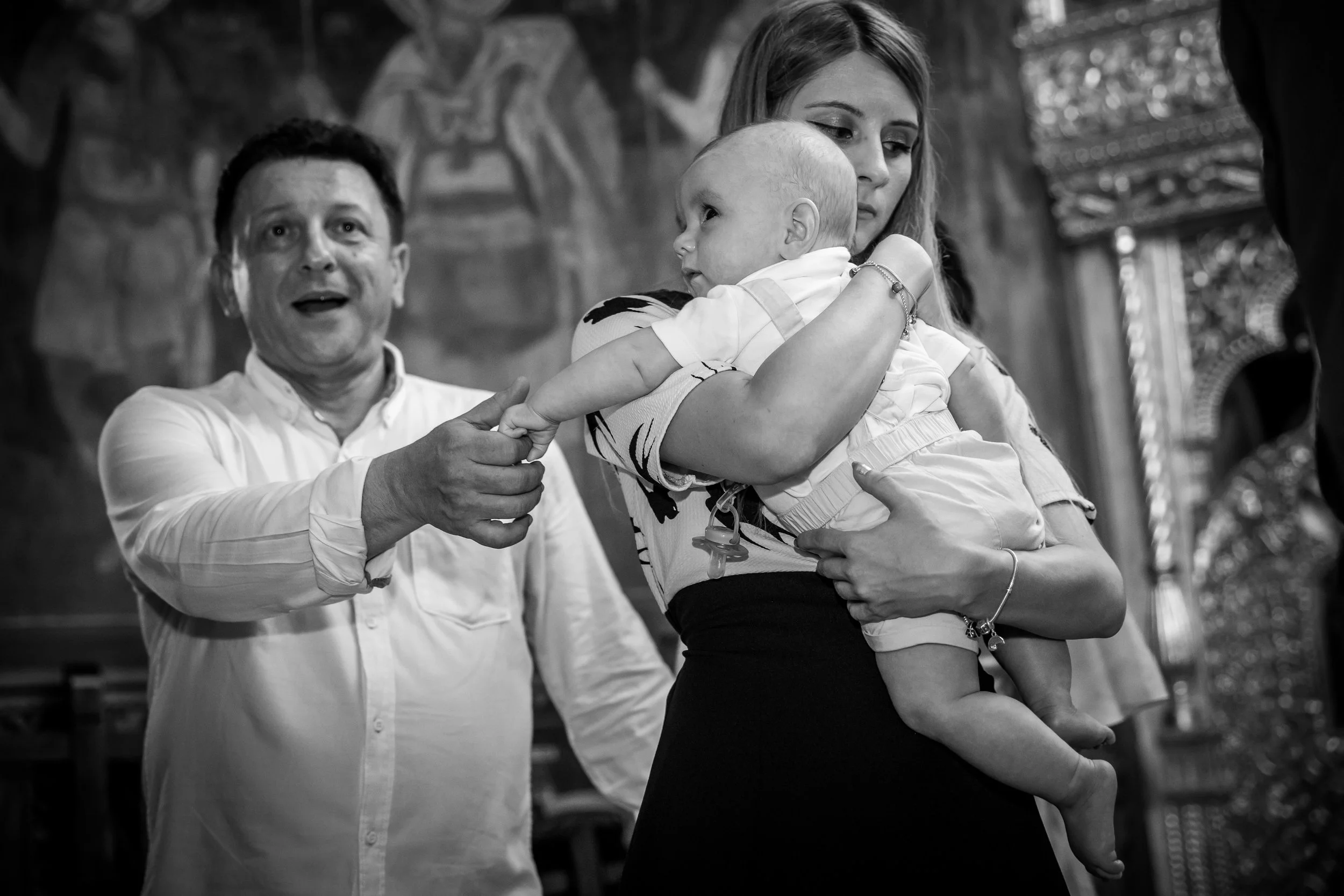 A black and white photo of a woman holding a baby, with a man smiling and looking at the camera in the background. The woman appears to be putting her hand on the baby's head, and the scene looks emotional, possibly from an event or celebration.