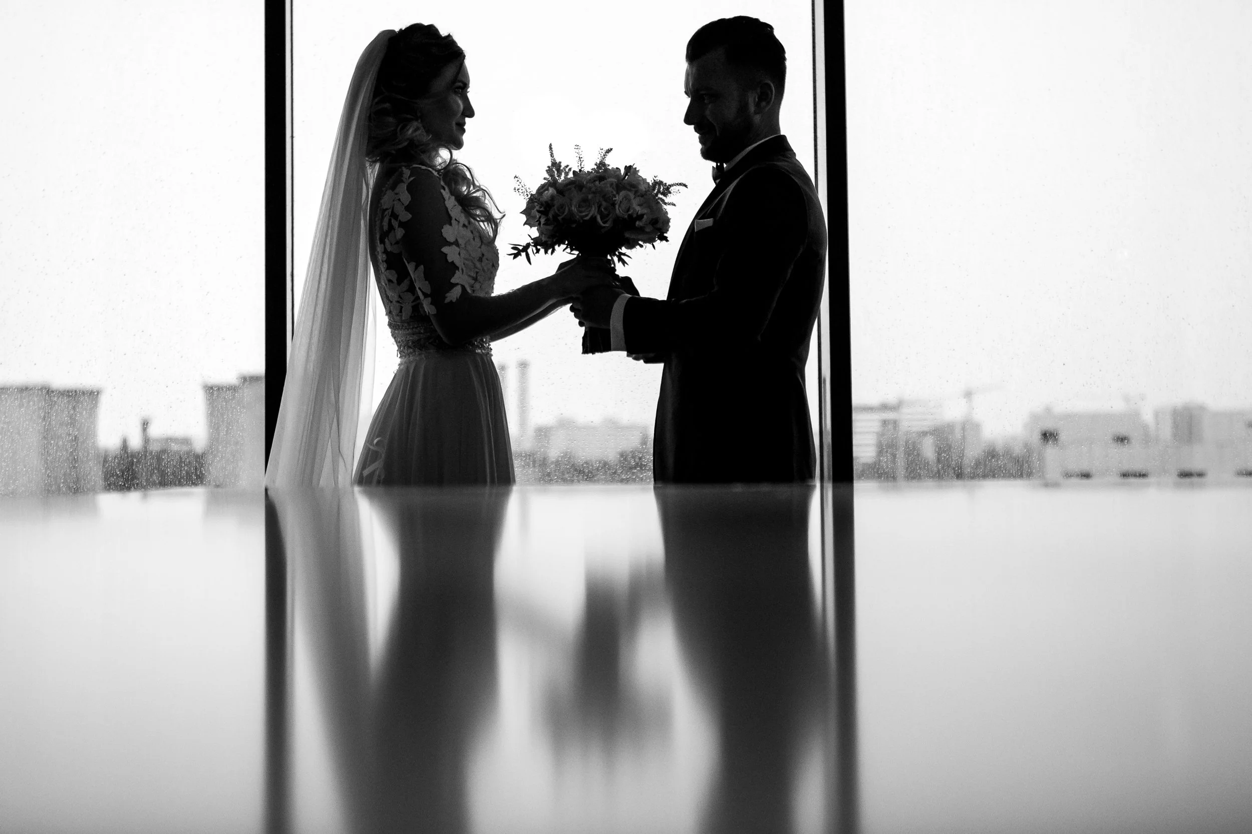 Silhouette of a bride and groom holding hands and exchanging a bouquet in front of a window with cityscape background.