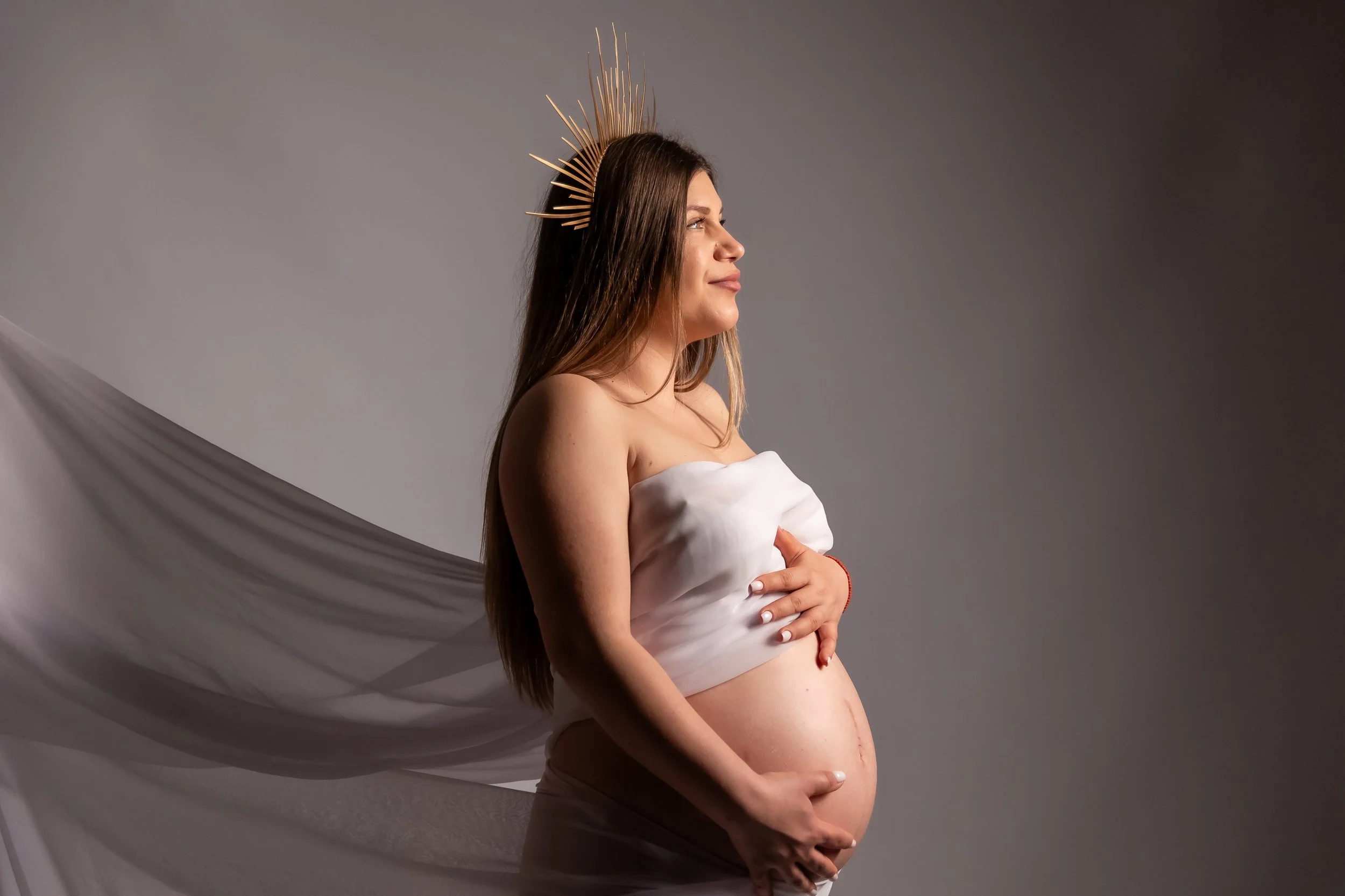 A woman with long brown hair and a halo-like headpiece made of wooden sticks, posing with her eyes closed and hand on her pregnant belly, wearing a white off-shoulder top, against a neutral gray backdrop with draped fabric.