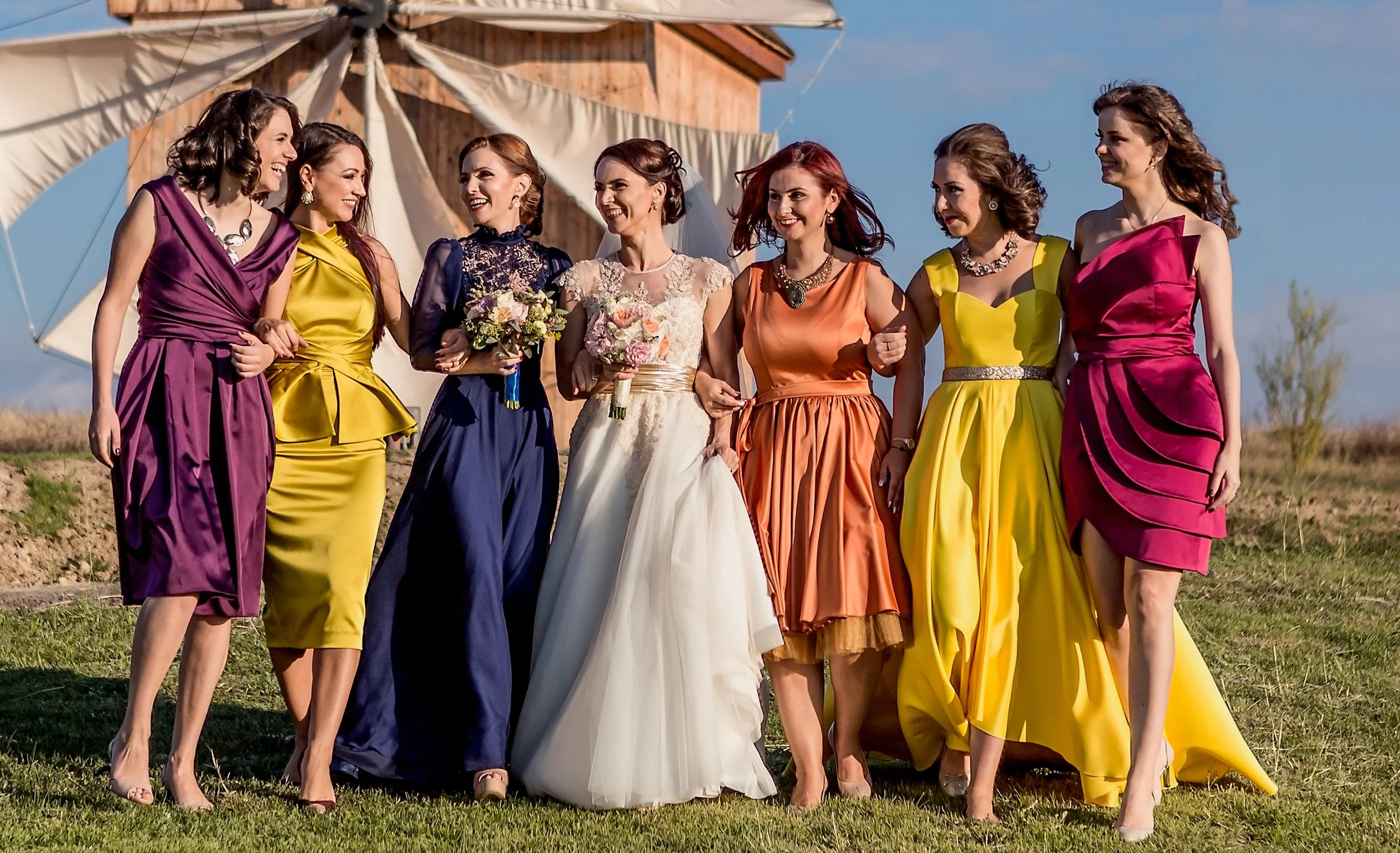 Group of women in colorful dresses walking outdoors with a wedding bride holding bouquets, smiling and enjoying a sunny day.
