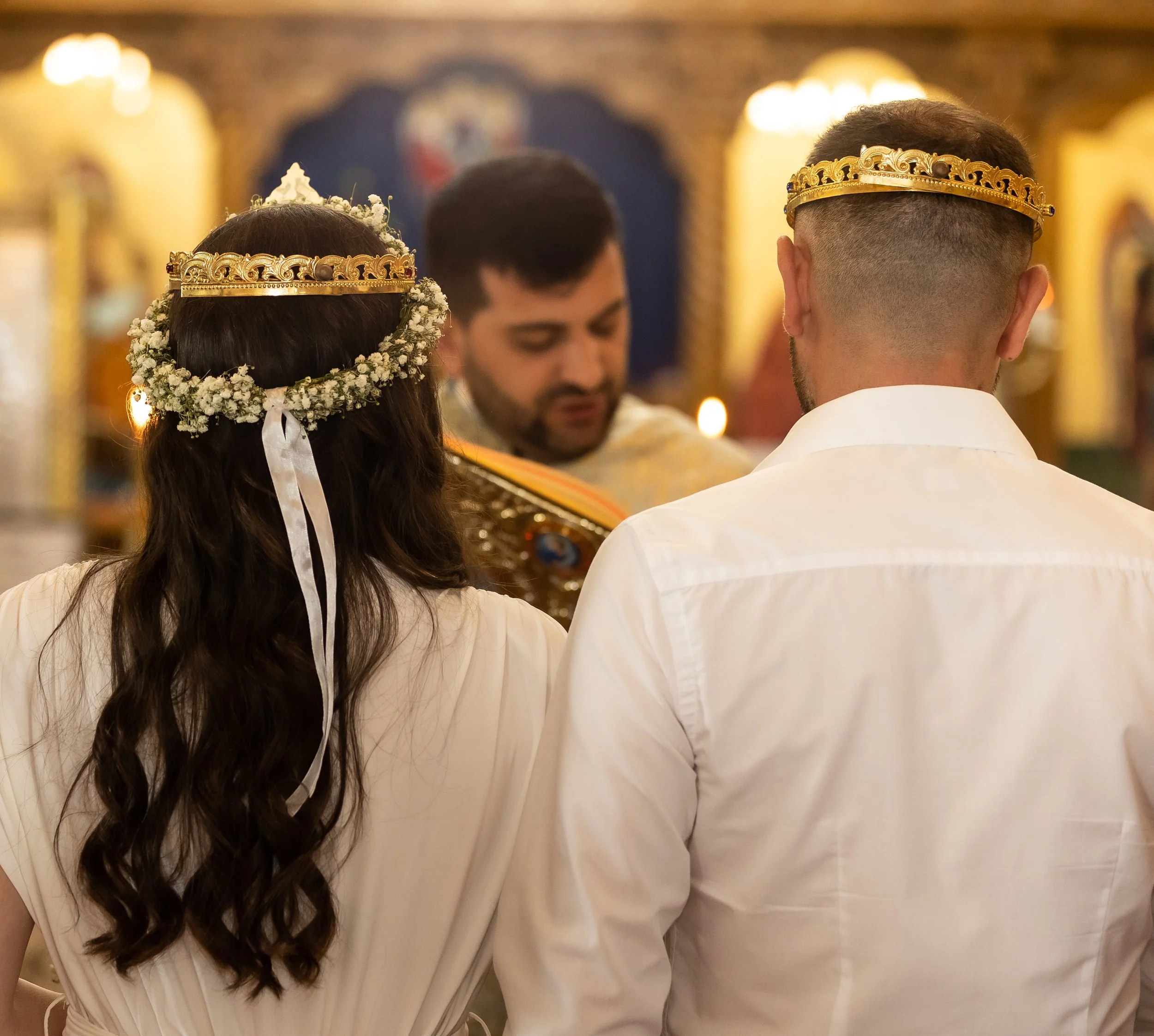 A woman and a man wearing crowns and white robes, possibly at a wedding or religious ceremony, standing in a decorated church or cathedral with ornate gold accents in the background.