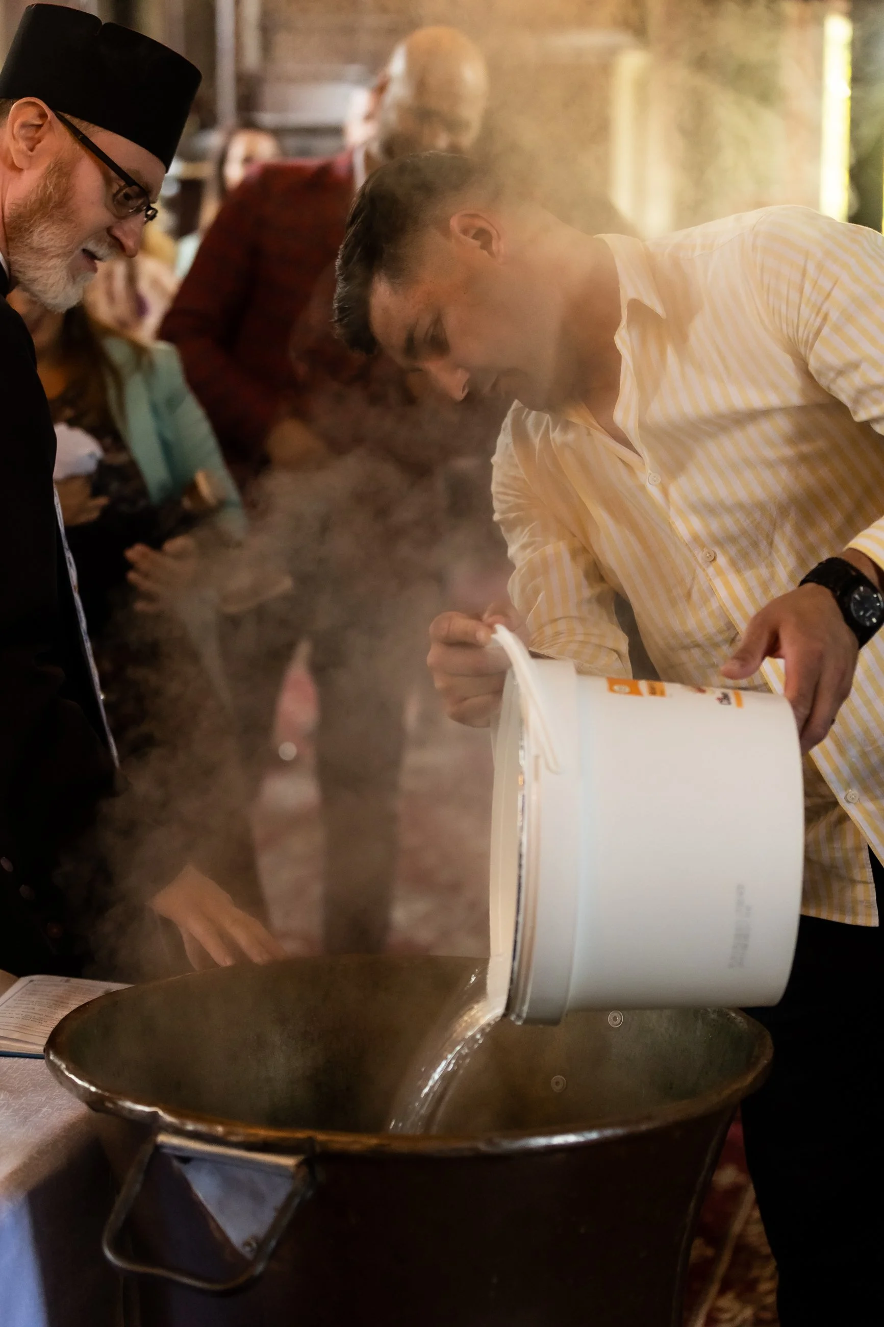 A chef cooking over a steaming pan in a restaurant kitchen, with a customer in a yellow checkered shirt looking at a menu or recipe book. Several people are in the background, observing and waiting.