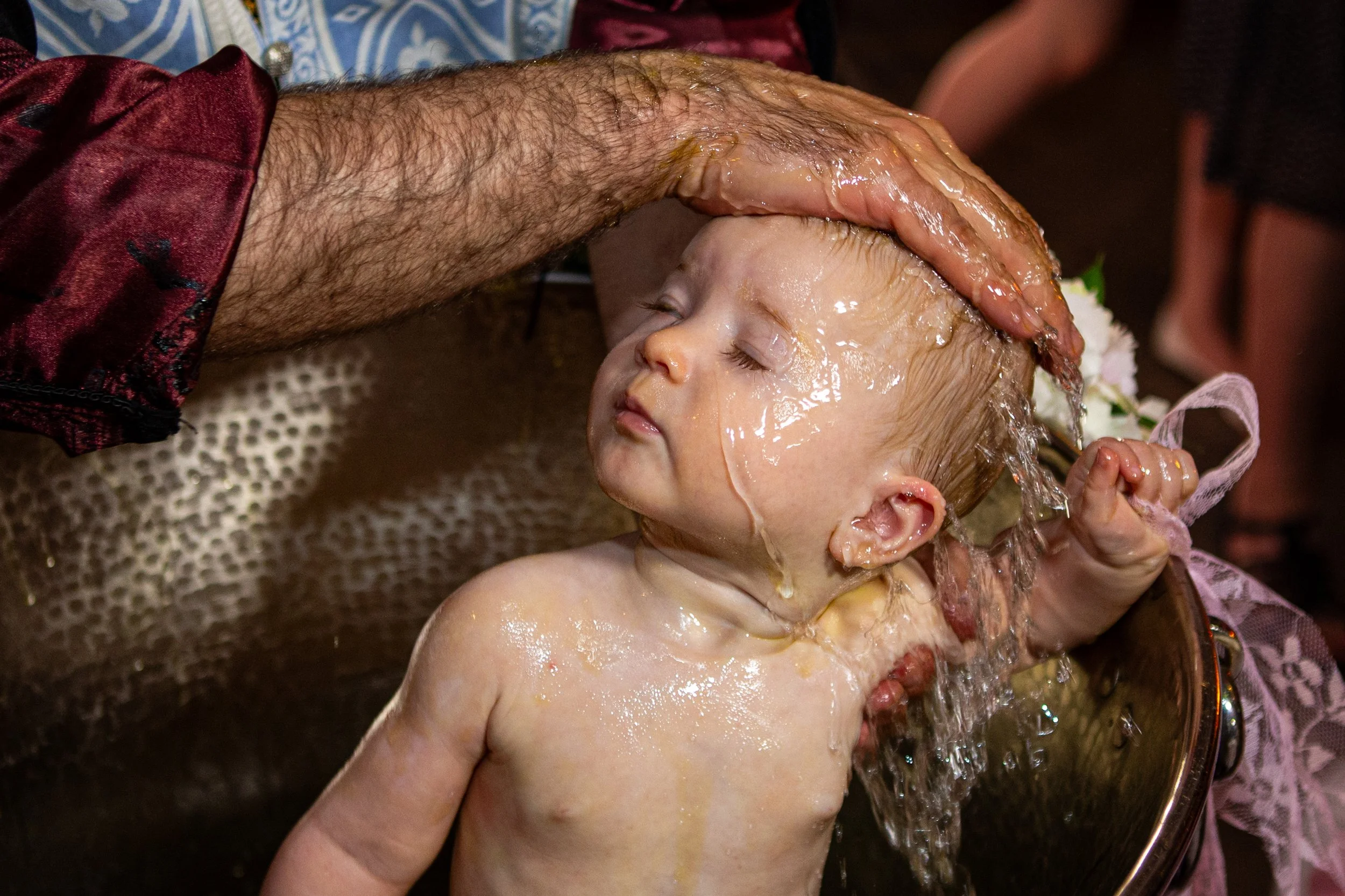 A young child with their eyes closed, receiving a traditional water cleansing ritual by an adult's hand pouring water over their head, with the child and adult partially submerged in a water basin.