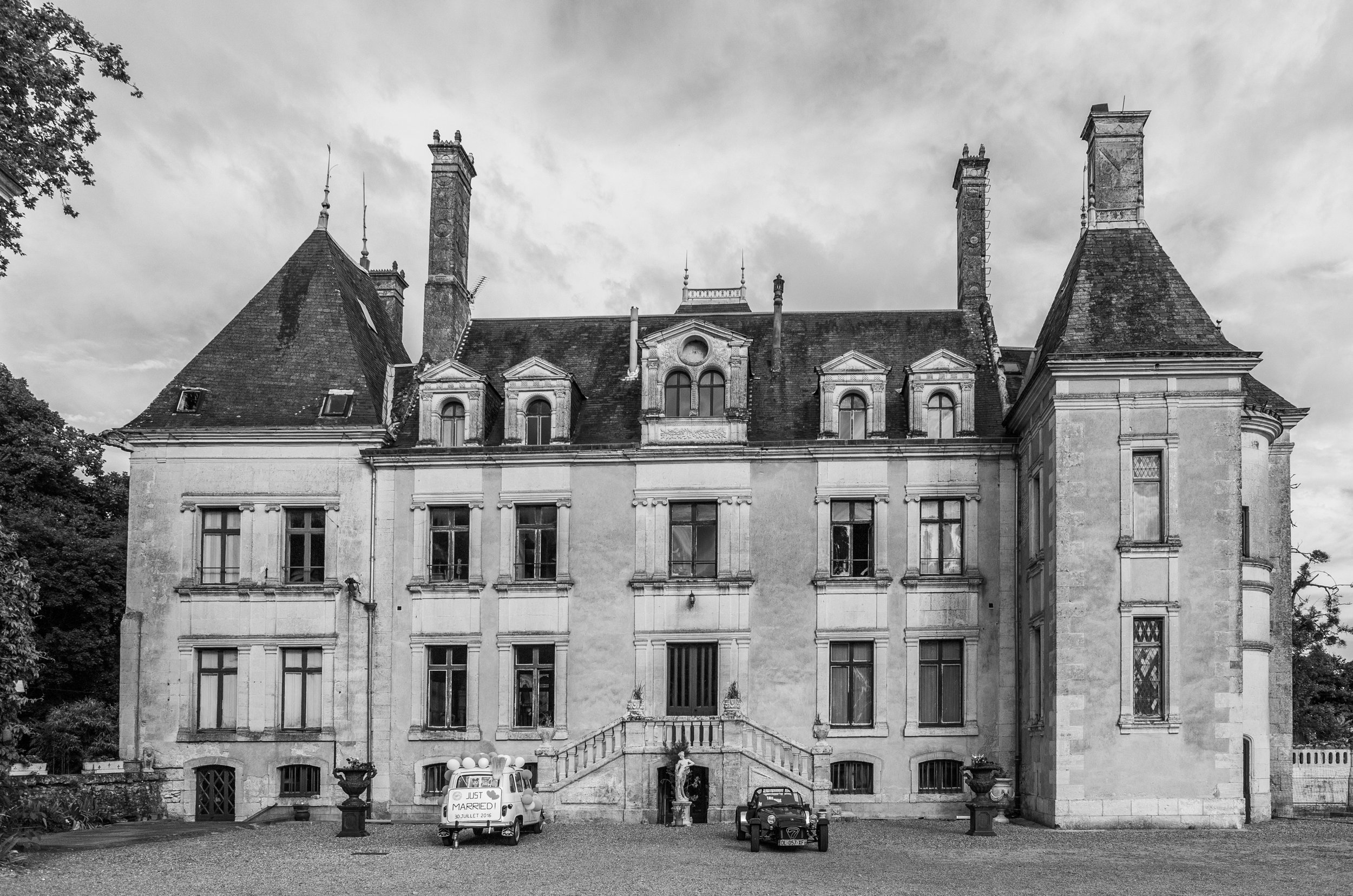 Black and white photo of a large historic mansion with tall chimneys, multiple windows, and decorative architectural features. In the front, there are a small vintage car with a sign that reads 'Just Married,' and a miniature car, along with decorati