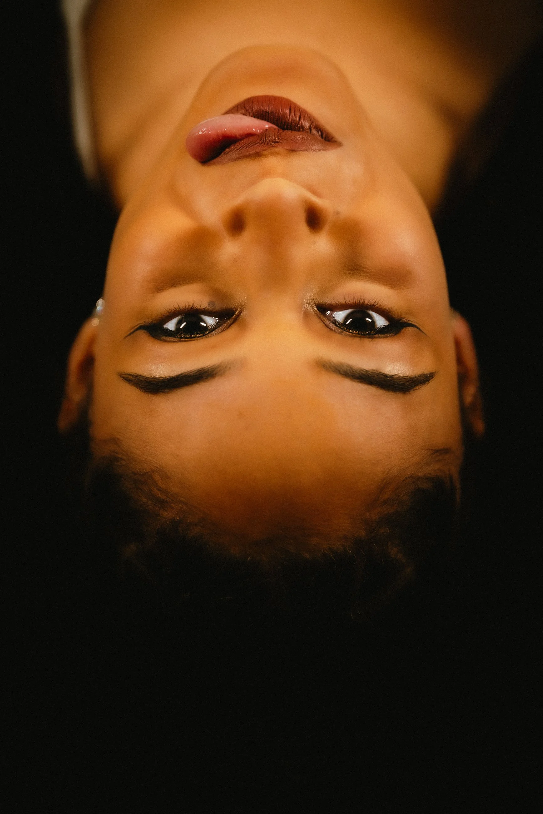 A person with light brown skin and full lips, lying down with head tilted back. The person has dark, curly hair, and is wearing eye makeup including dark eyeliner and mascara. The person's tongue is sticking out slightly, and they have ear piercings 