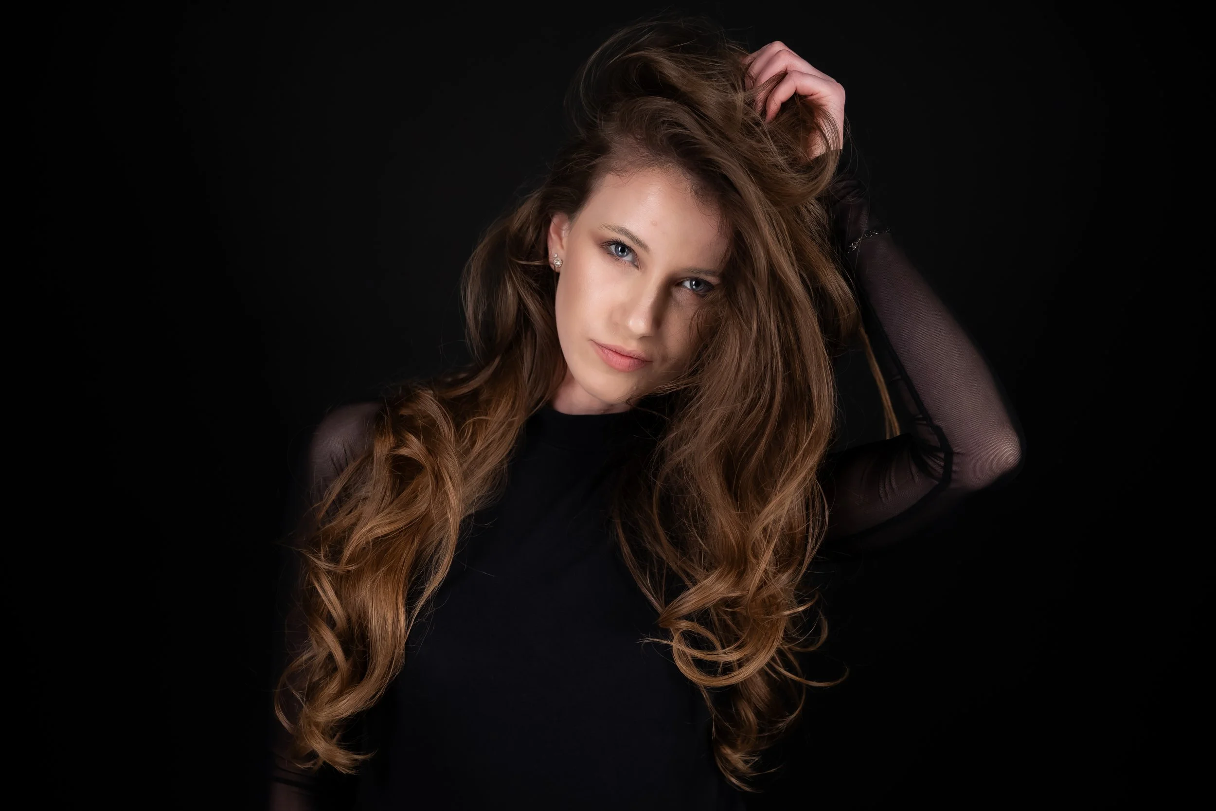 A woman with long, wavy brown hair, wearing earrings, poses against a black background, holding her hair back with one hand and looking at the camera.