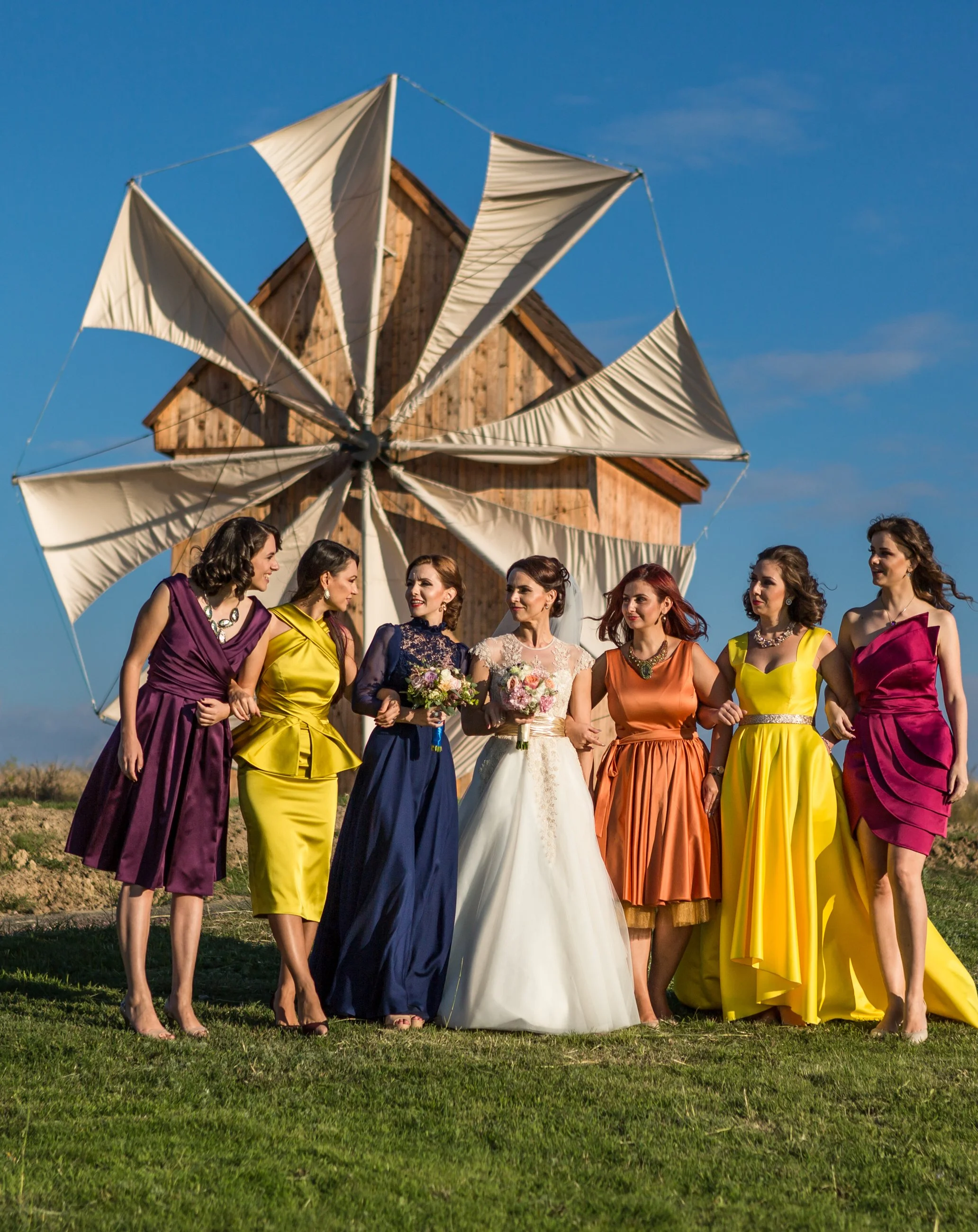 Women in colorful dresses standing outdoors on grass with a wooden windmill and a clear blue sky in the background.