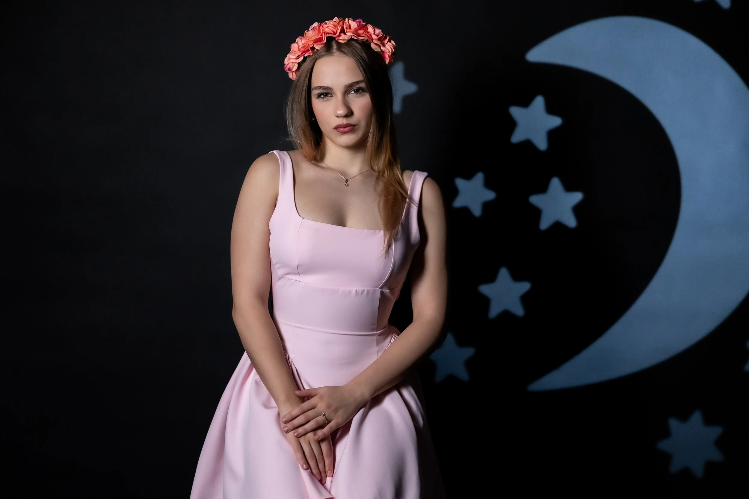 A young woman wearing a pink dress and flower crown stands against a black background with star-shaped shadow projection.