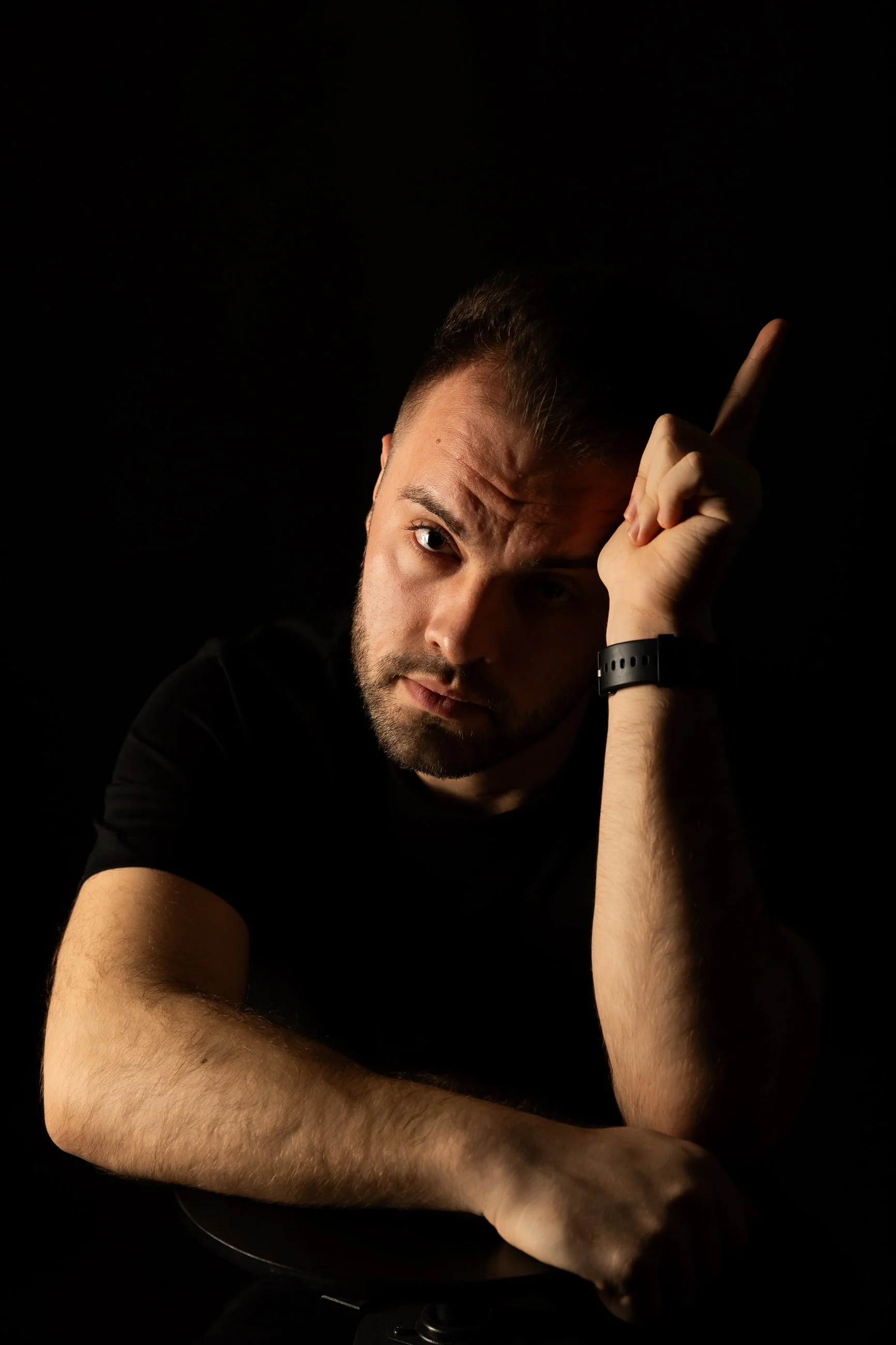 A man with short hair and a beard pointing upward with his index finger, resting his head on his hand, against a black background.