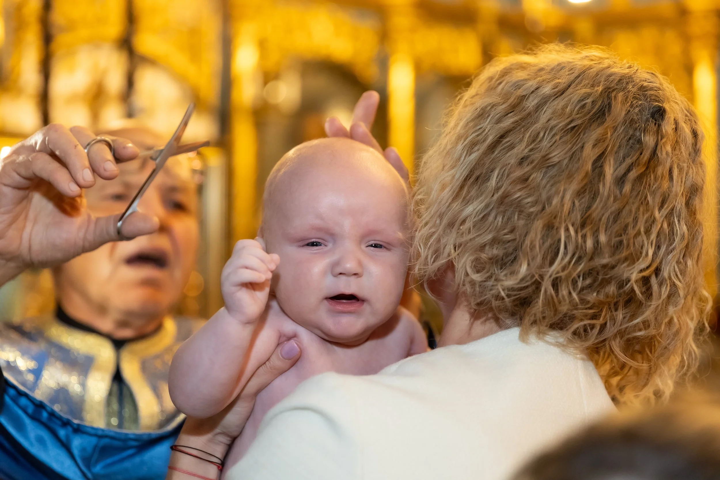A woman holding a baby with curly hair, and an older woman in the background, under warm yellow lighting.