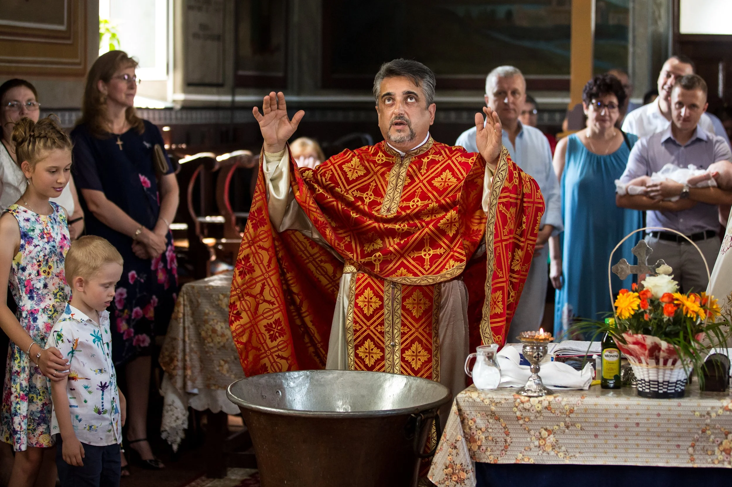 A priest dressed in ornate red and gold liturgical vestments stands at the center of a religious ceremony, with his arms raised and eyes looking upward. He is surrounded by a diverse group of people, including children and adults, inside a church dec