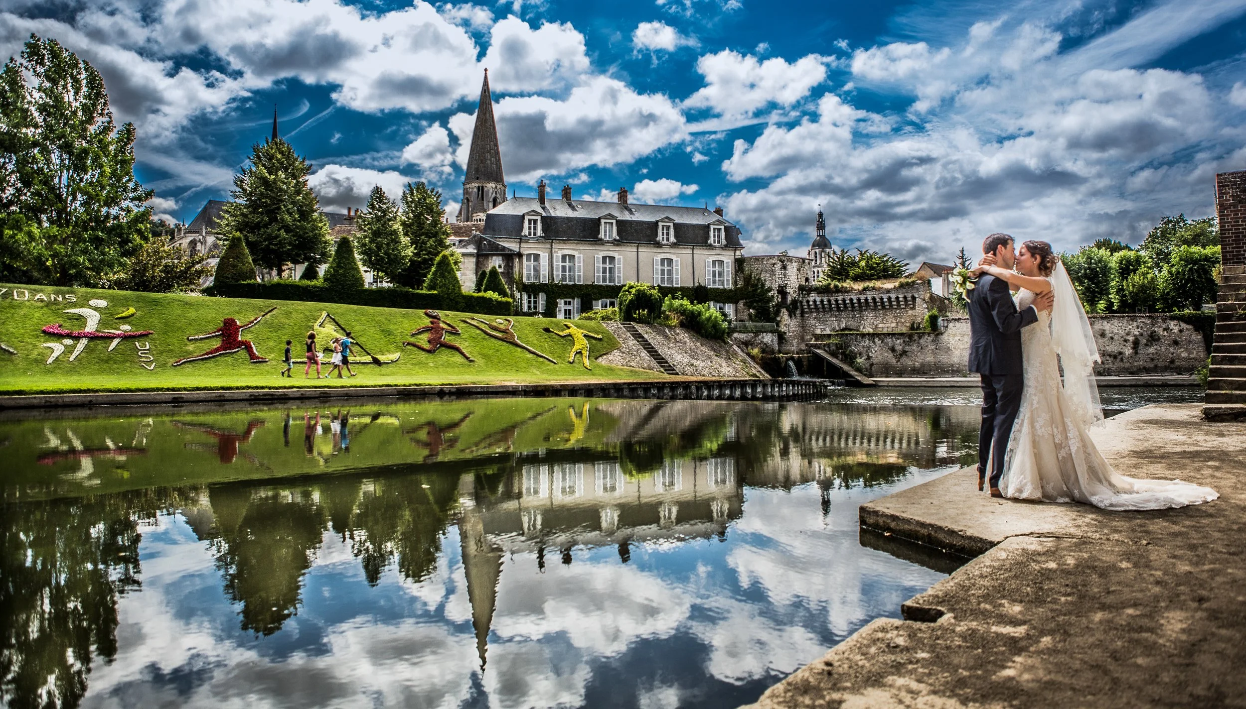 A bride and groom embrace near a river on a cloudy day, with historic buildings and a church steeple in the background.