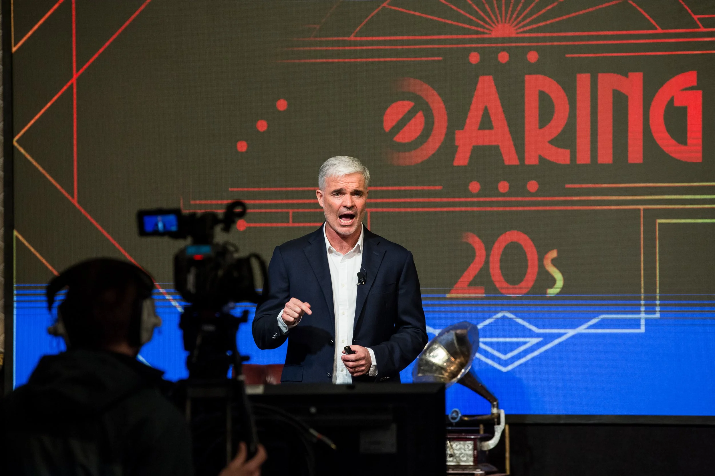 A man with gray hair and a navy suit giving a speech or presentation in front of a large screen displaying the word 'DARLING' and some numbers. There are cameras and audience members in the foreground.