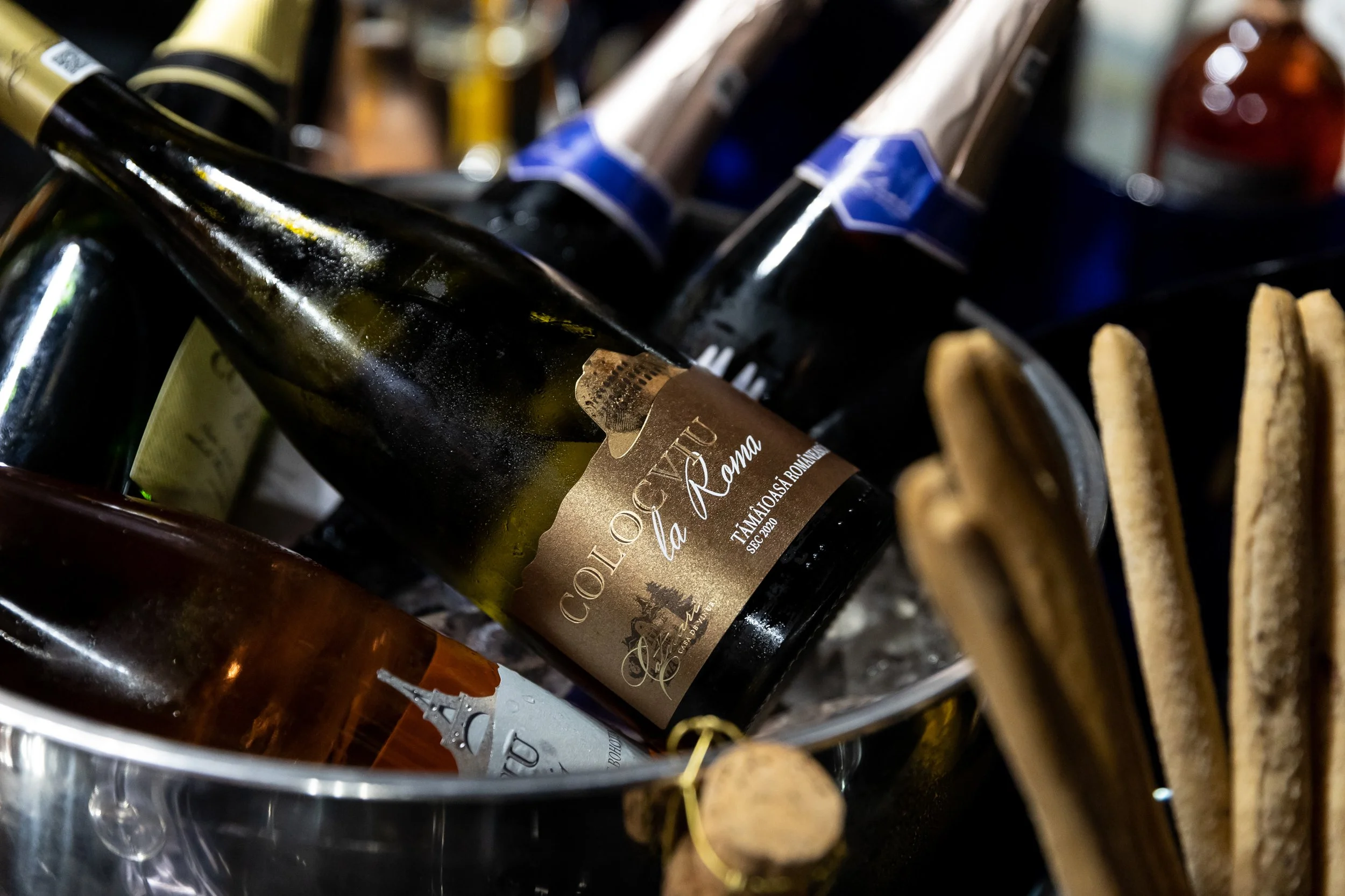 Close-up of wine bottles in a wine rack, with one bottle prominently labeled 'Cea de la Roma' and others with varied labels, some with foil capsules, showing wine stored for aging or display.