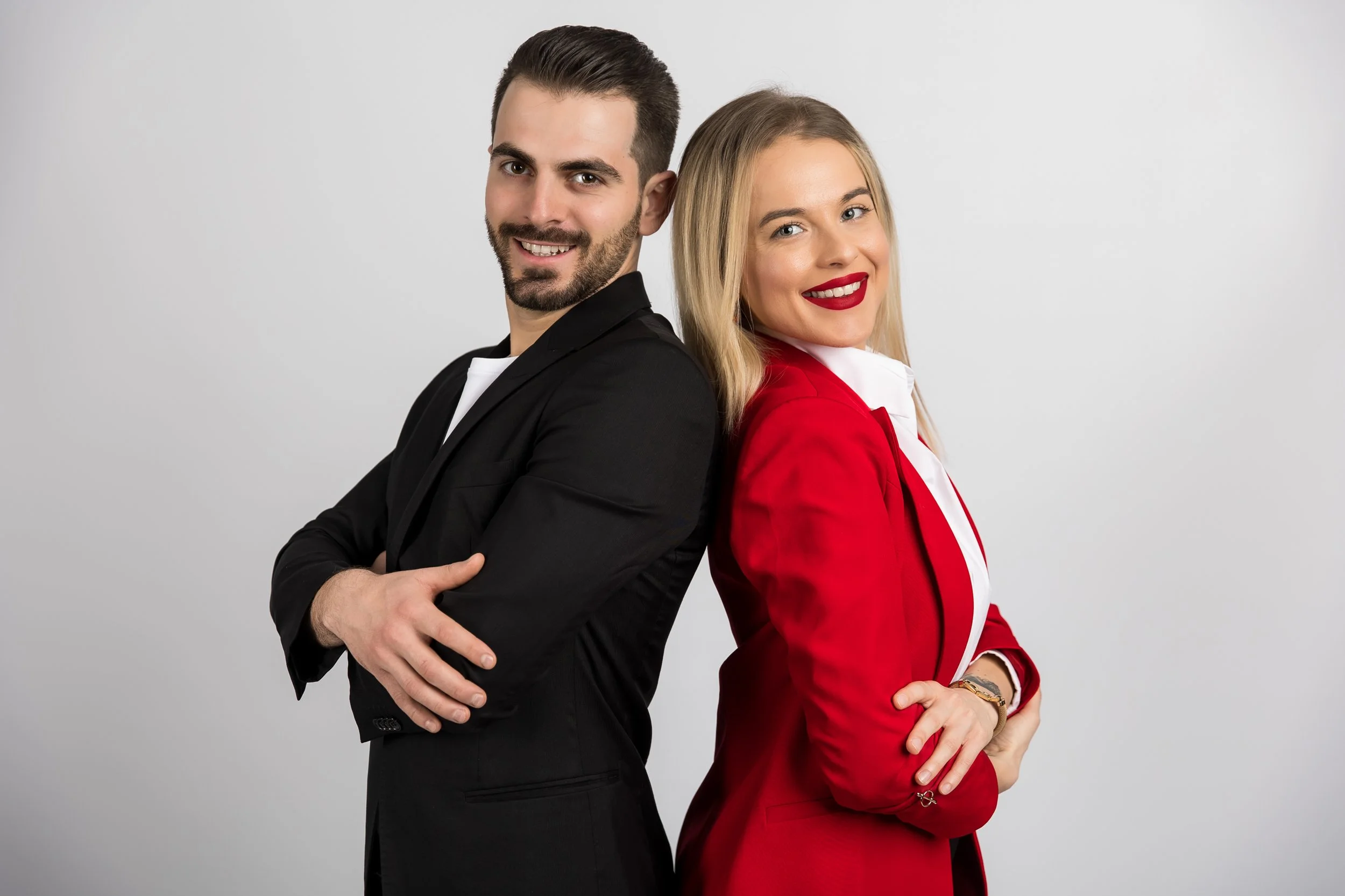 A man and woman stand back-to-back with arms crossed, smiling, against a plain white background.