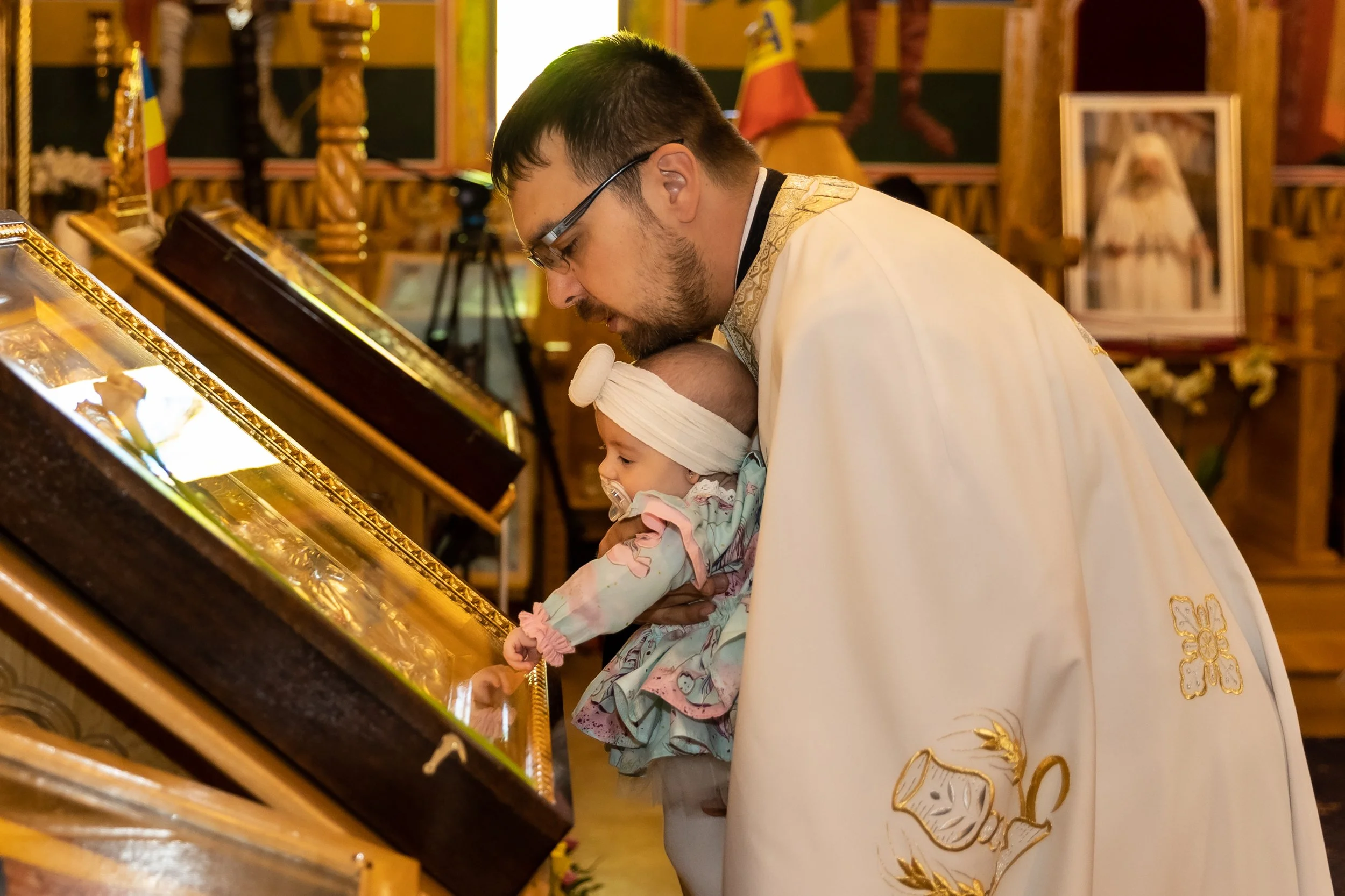 A priest wearing white robes with gold embroidery is holding a baby dressed in patterned clothes and a headband, as they both look at a religious display inside a church.