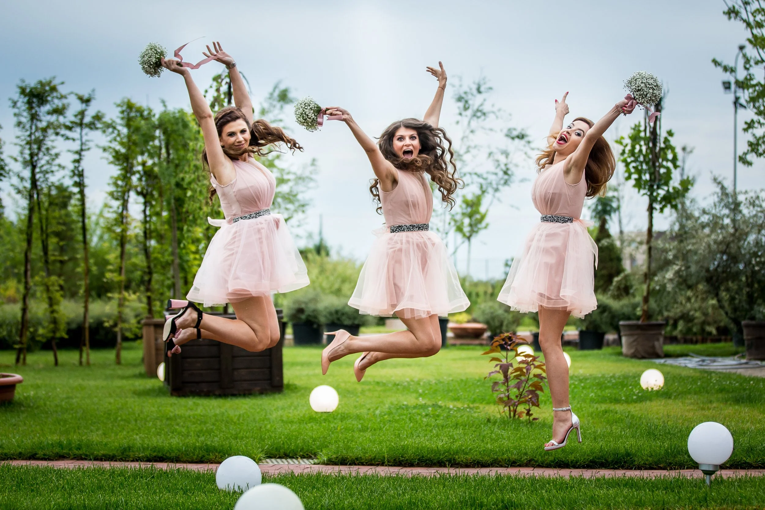 Three women in pink dresses jumping in a garden, holding small bouquets, with trees and outdoor lights in the background.