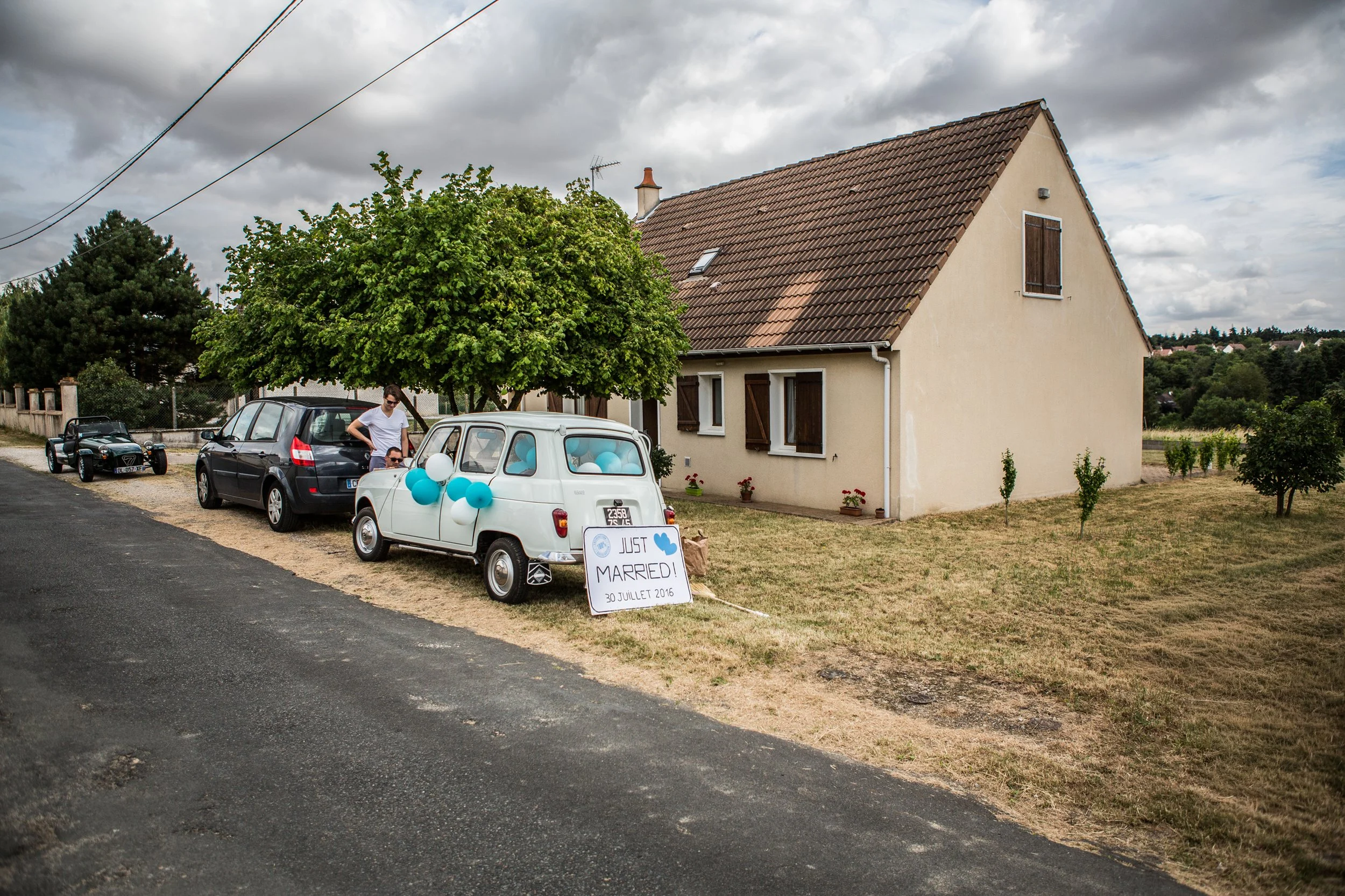 A small white car decorated with blue and white balloons is parked on a grassy lawn in front of a house. There is a sign that reads 'Just Married! 30 Juillet 2016' leaning against the car, indicating a wedding celebration. A man is standing next to t