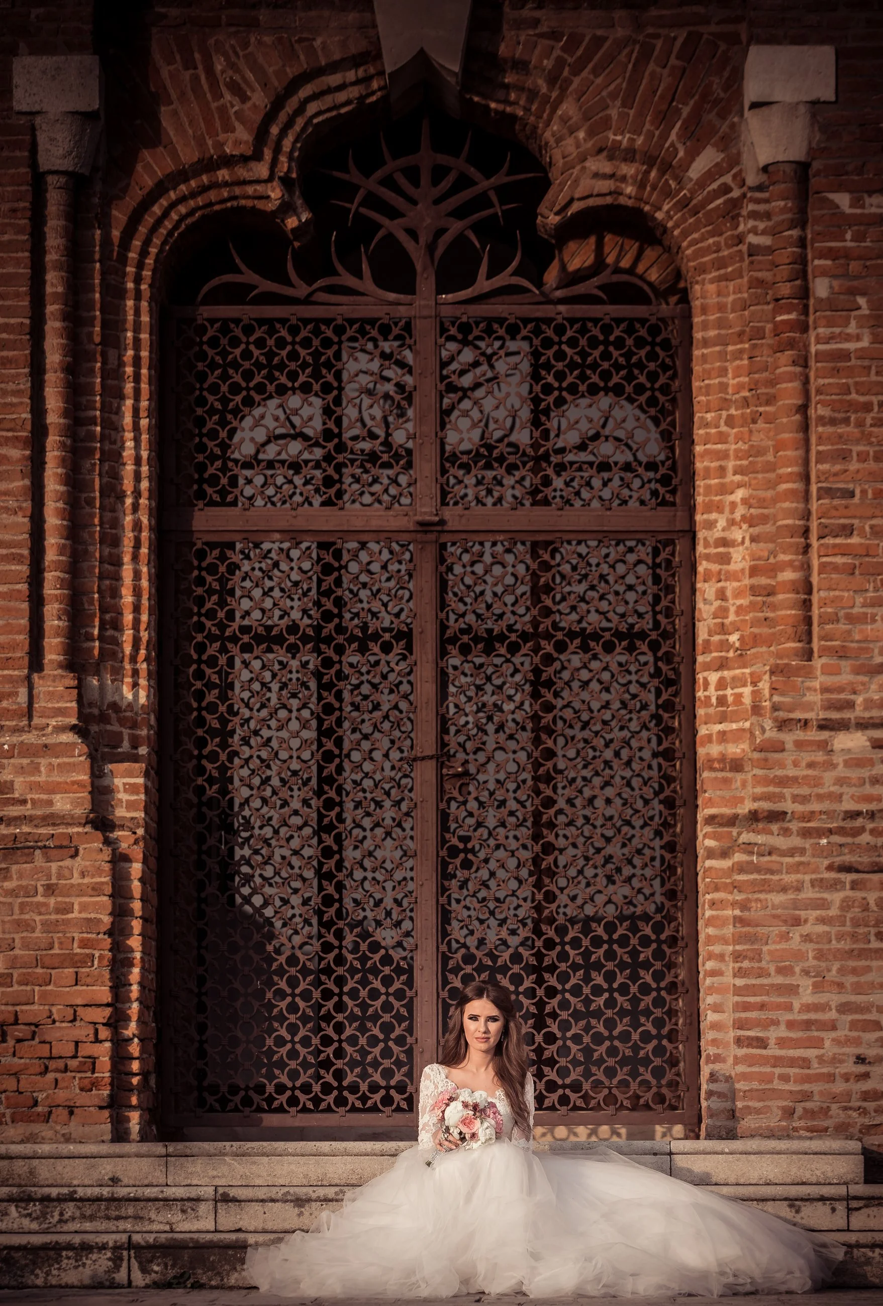 A bride sitting on stone steps in front of an ornate metal gate with brick walls on either side, holding a bouquet of flowers, wearing a white wedding dress with a long, flowing tulle train, and looking at the camera.