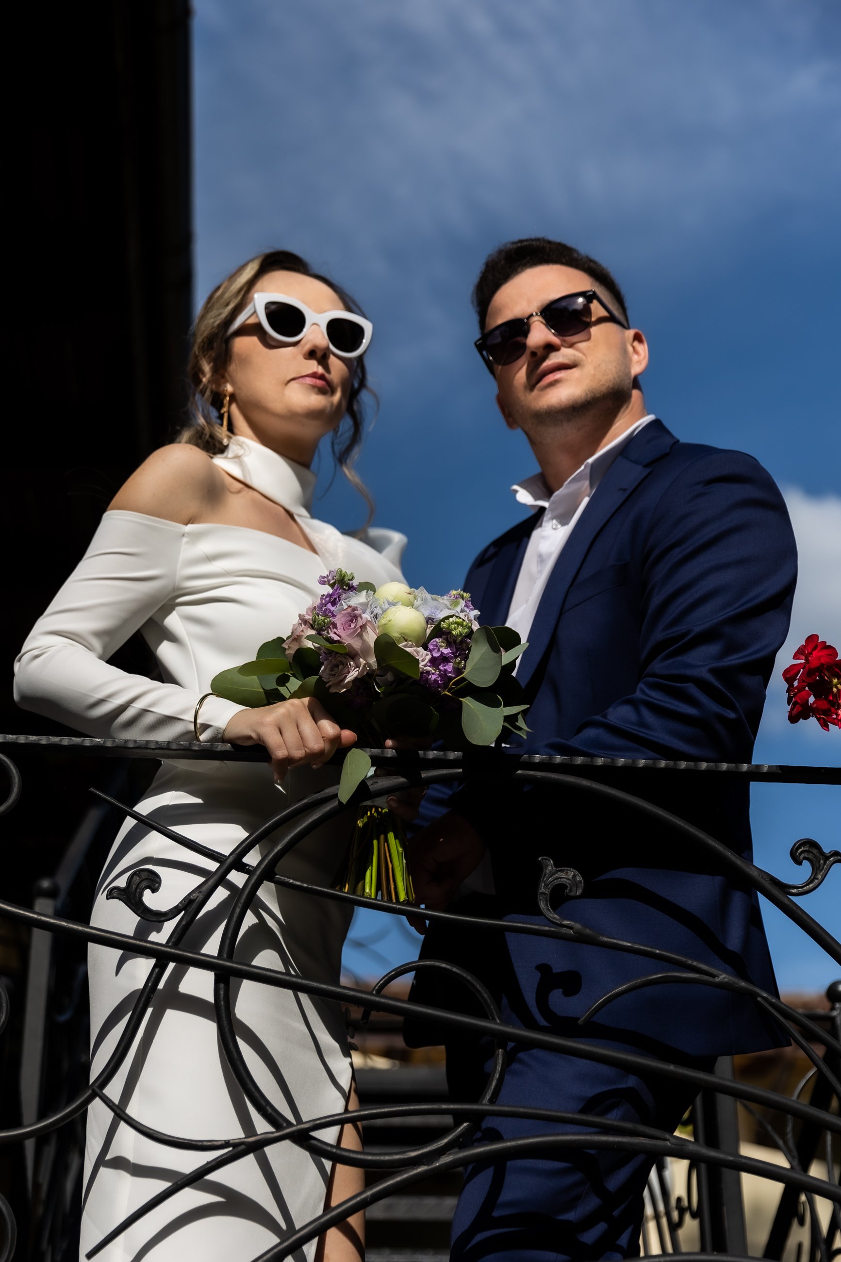 A man and woman wearing sunglasses and formal attire standing close together outdoors, holding a bouquet of flowers, with a blue sky in the background.