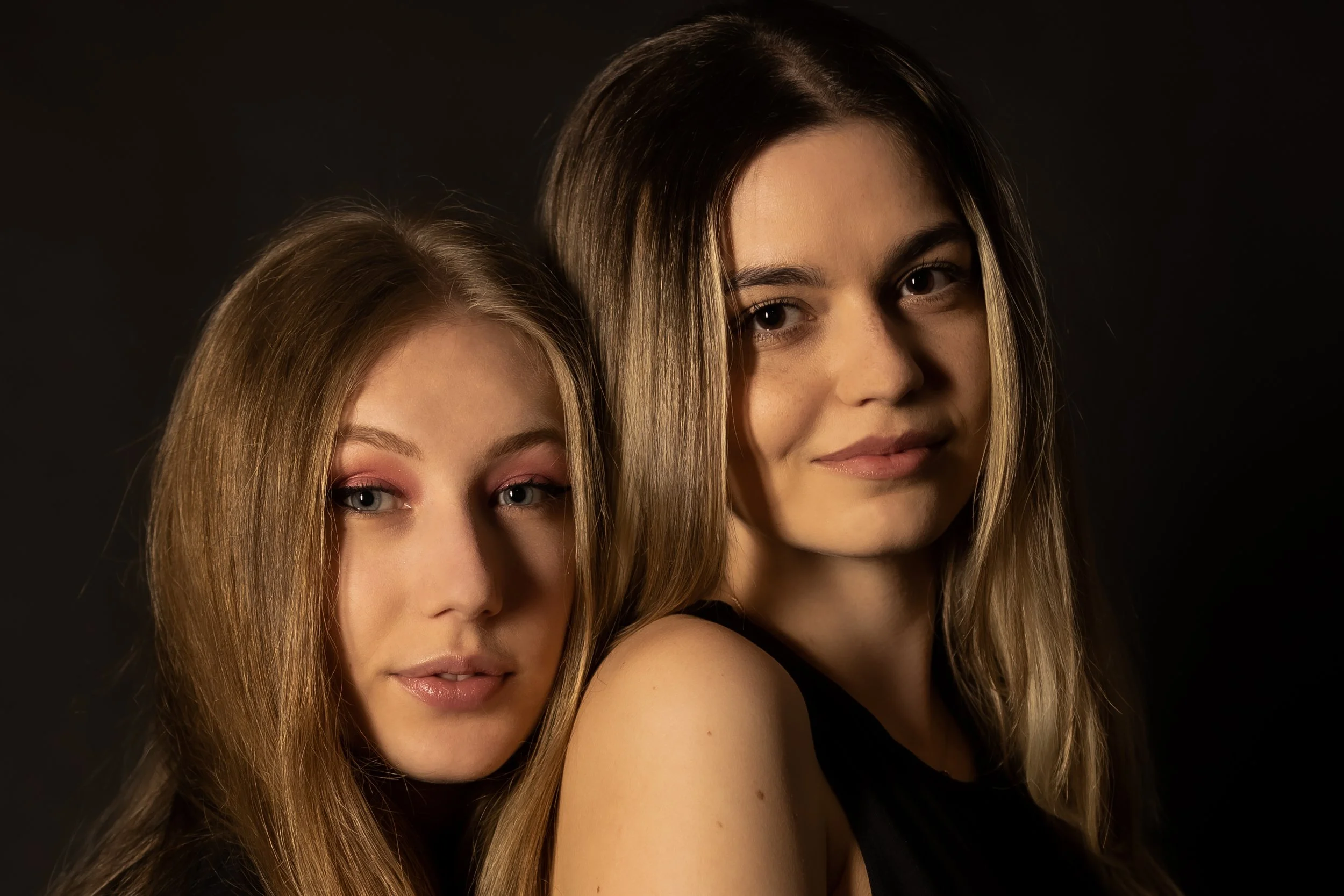 Close-up portrait of two young women with long wavy hair, minimal makeup, and black outfits against a dark background.