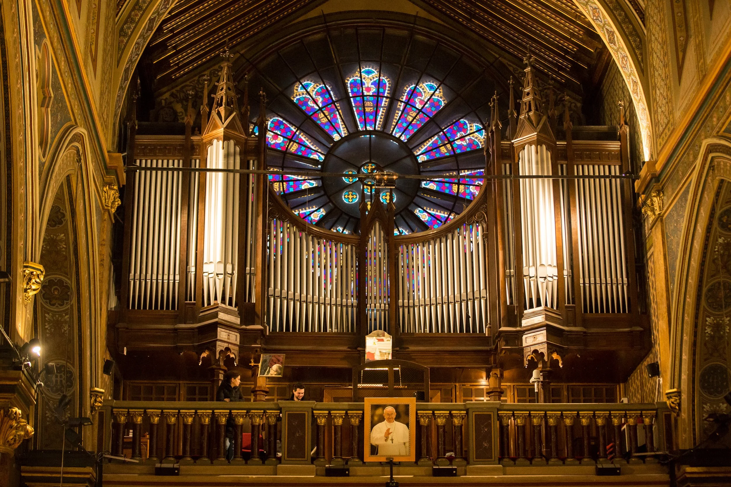 Interior of a church or cathedral featuring a large, ornate pipe organ with stained glass windows above. There are people near the front, and the setting appears formal and historic.