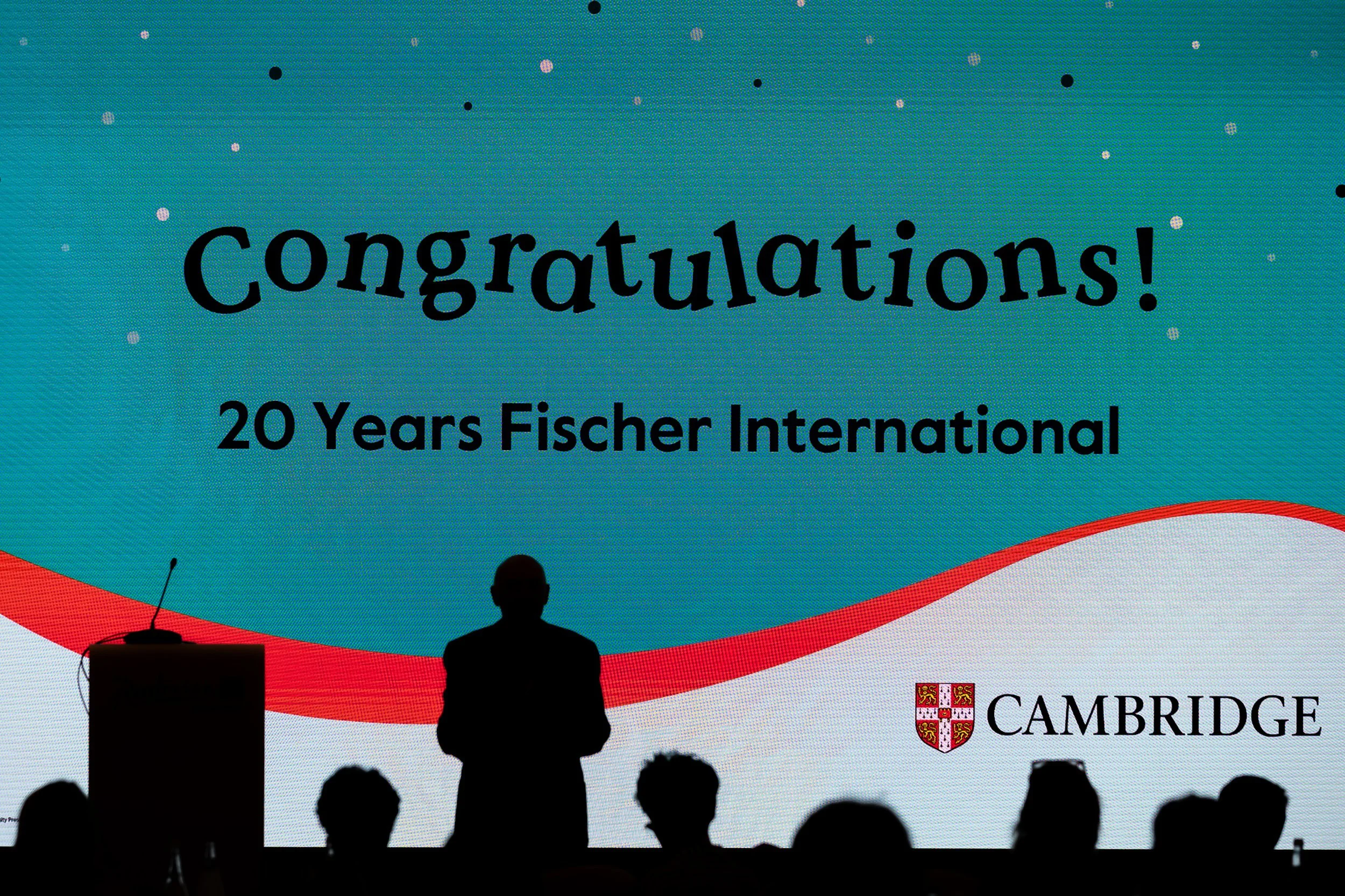A large screen displaying 'congratulations!' and '20 Years Fischer International,' with a silhouette of a person speaking at a podium and an audience in front, with the Cambridge shield logo in the bottom right corner.