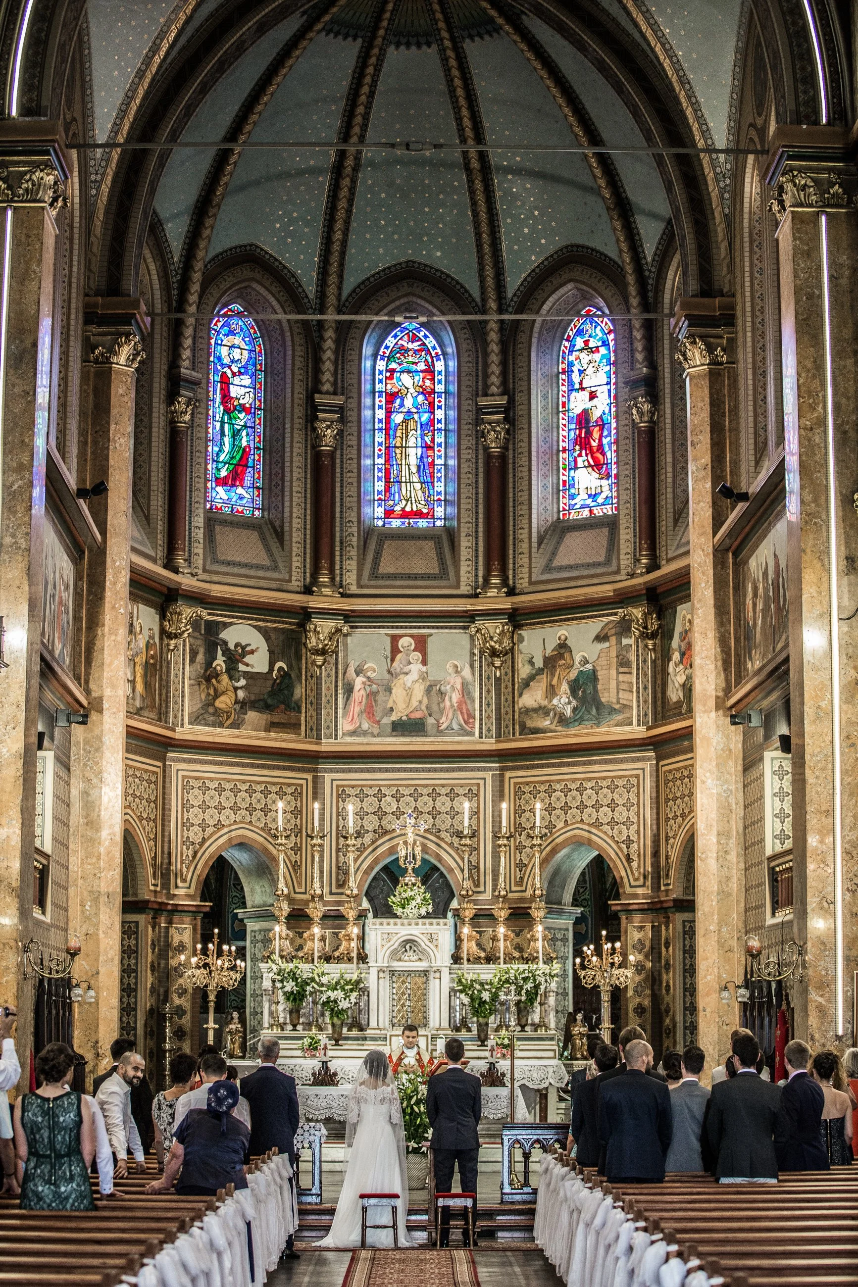 A wedding ceremony inside a church with a bride and groom standing before the altar, surrounded by guests and ornate stained-glass windows in the background.