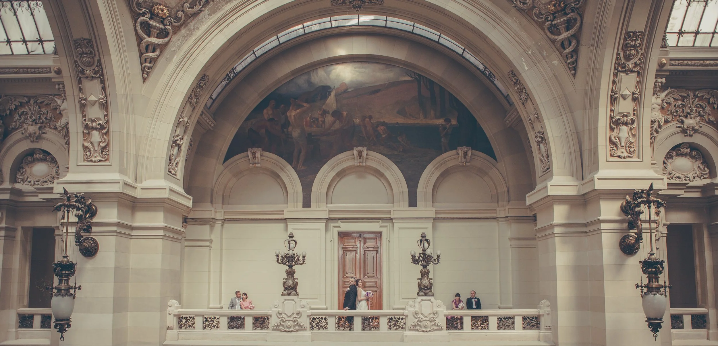 Elegant interior of a historic building with ornate architectural details, including carvings and a large mural of a rural scene, with a bride and groom standing on a balcony and a few guests seated nearby.