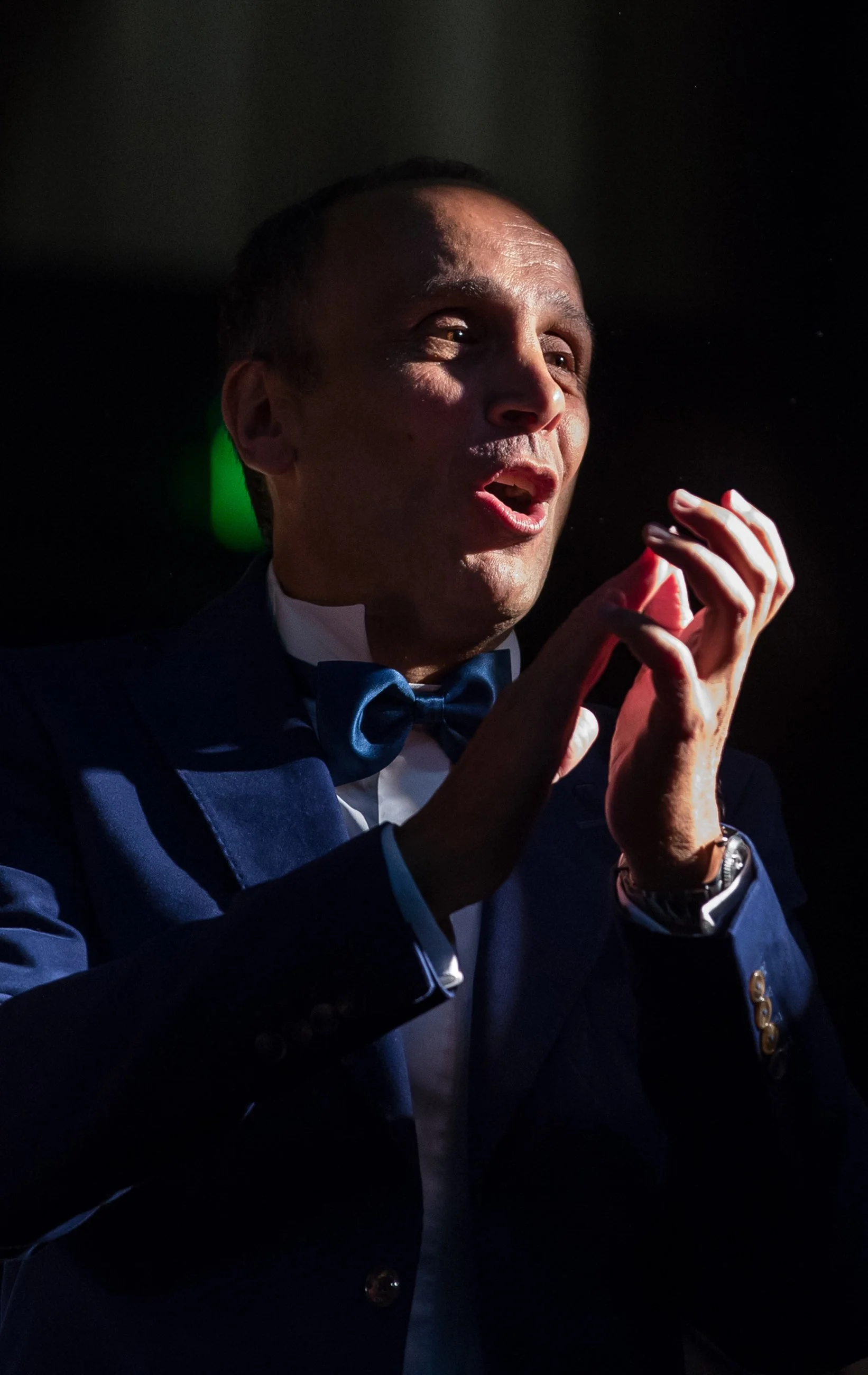 A man in a tuxedo with a bow tie claps his hands, with dramatic lighting highlighting his face against a dark background.