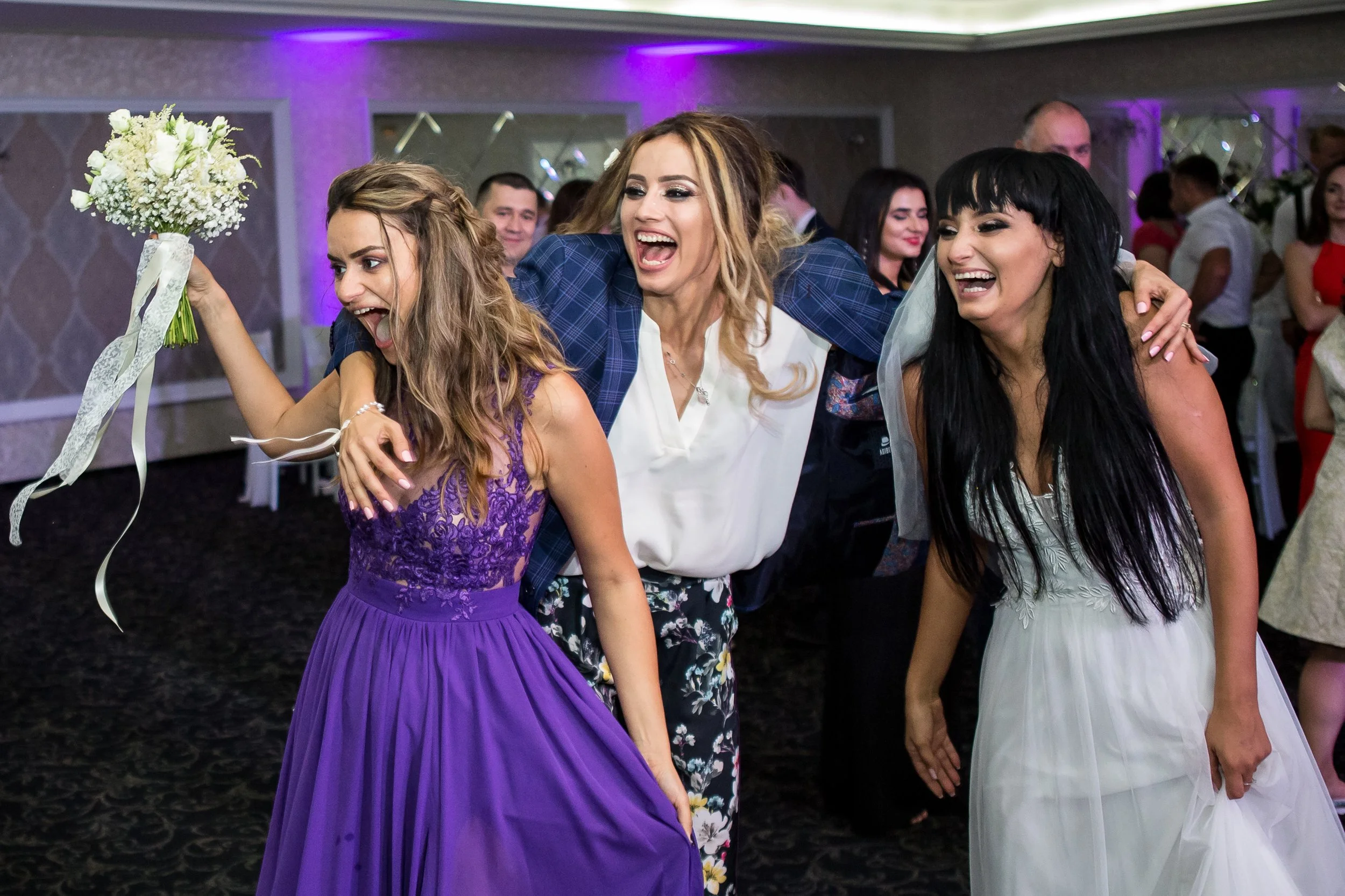 Three women are celebrating at a wedding reception, smiling, holding hands, and wearing dresses, with flowers and guests in the background.