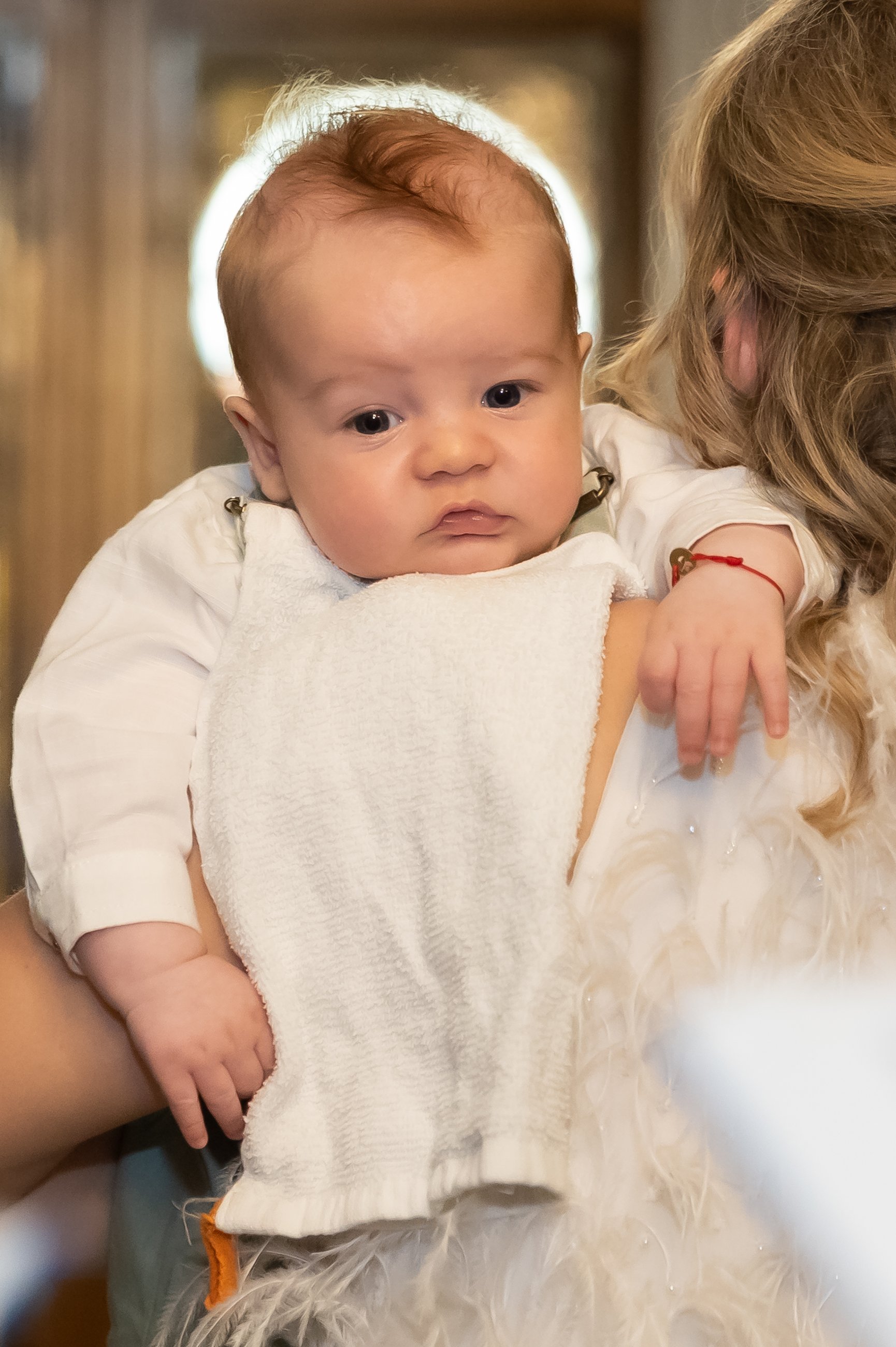 Close-up of a young baby with light skin and reddish-brown hair, being held by a person with blond hair, against a blurred background with a circular light. The baby is wearing a white outfit and a bib.