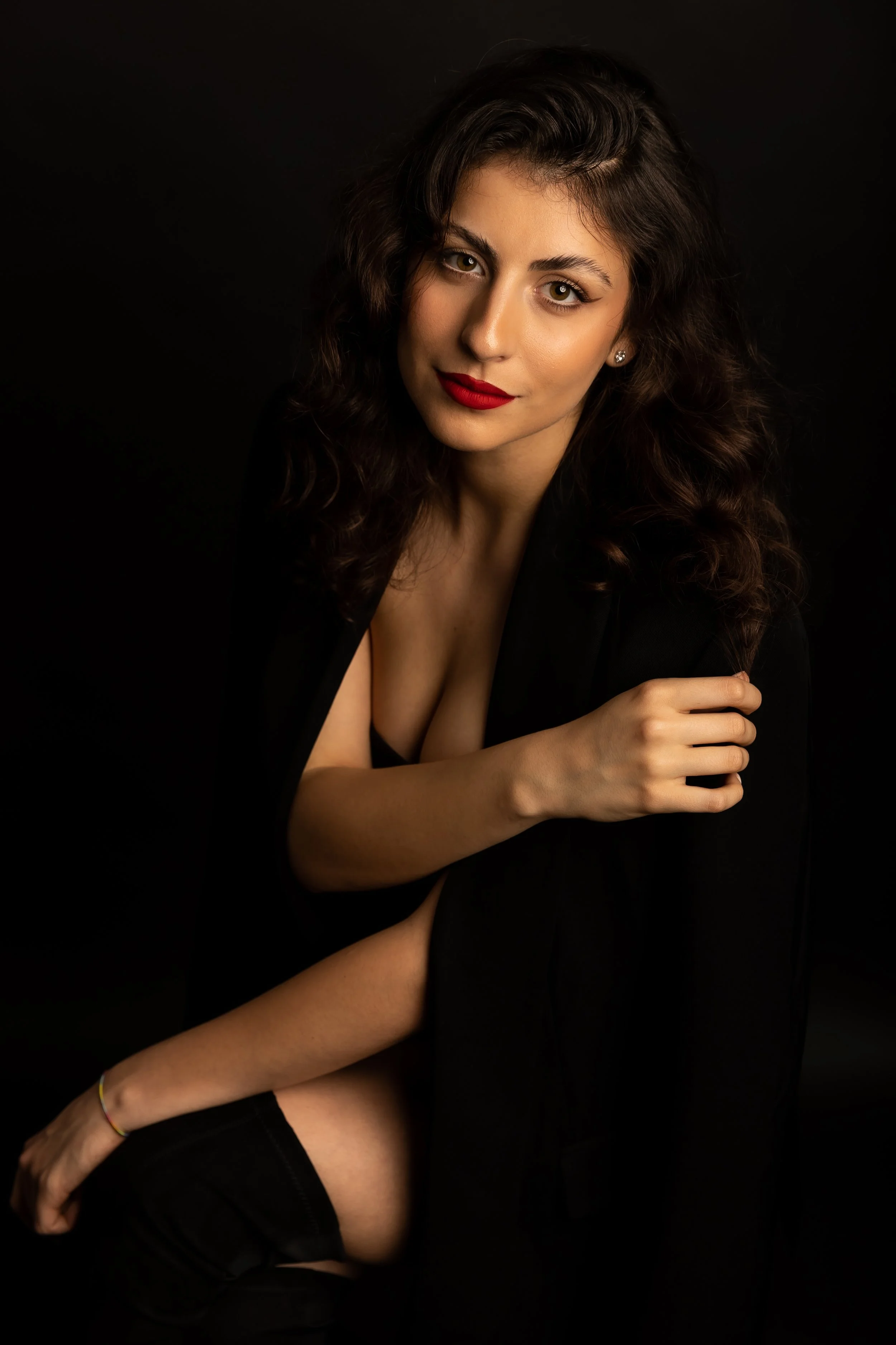 A woman with wavy dark hair, red lipstick, and earrings, posing against a black background.