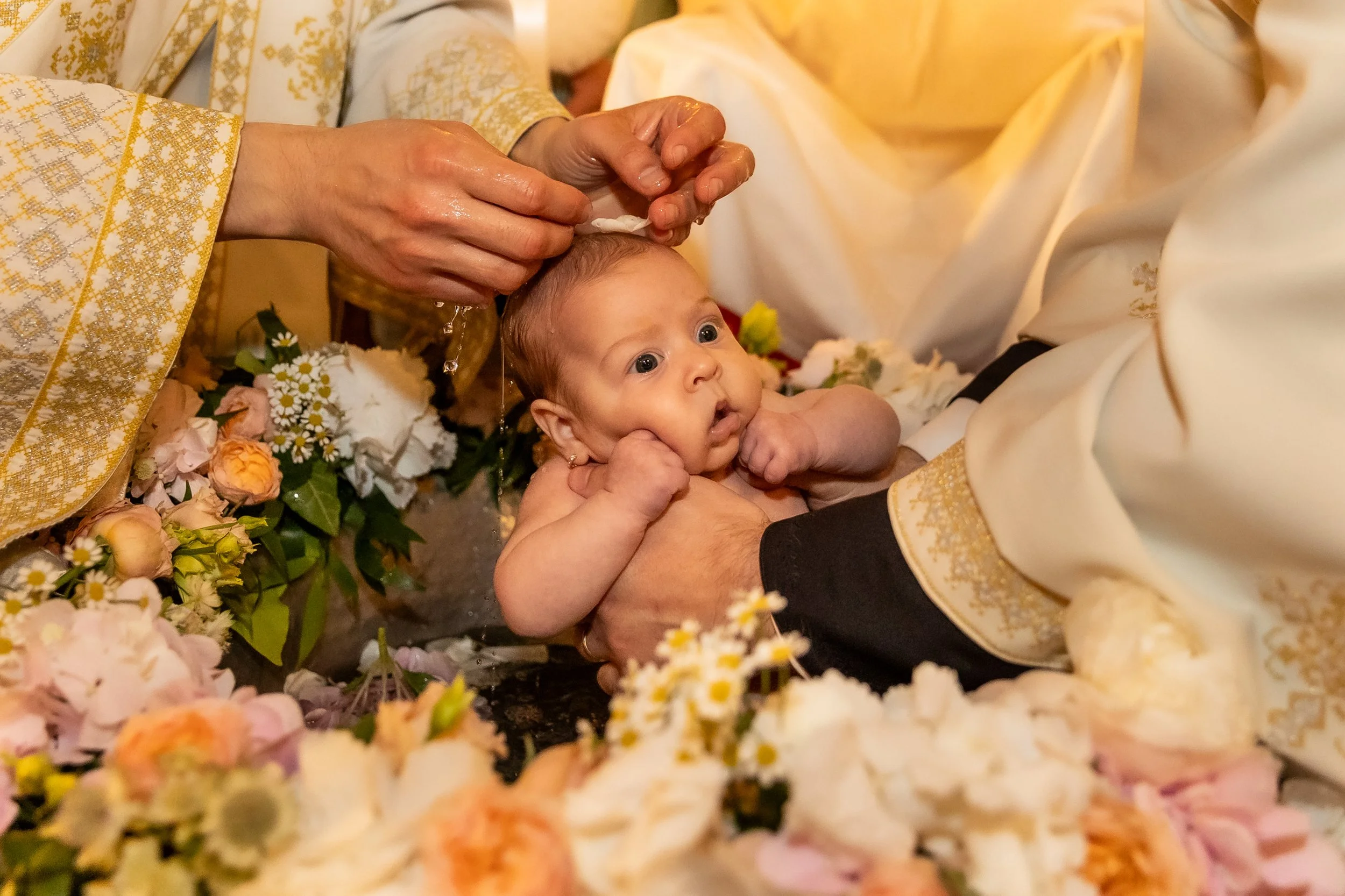 A baby being baptized by a priest dressed in ornate robes during a religious ceremony, surrounded by flowers.